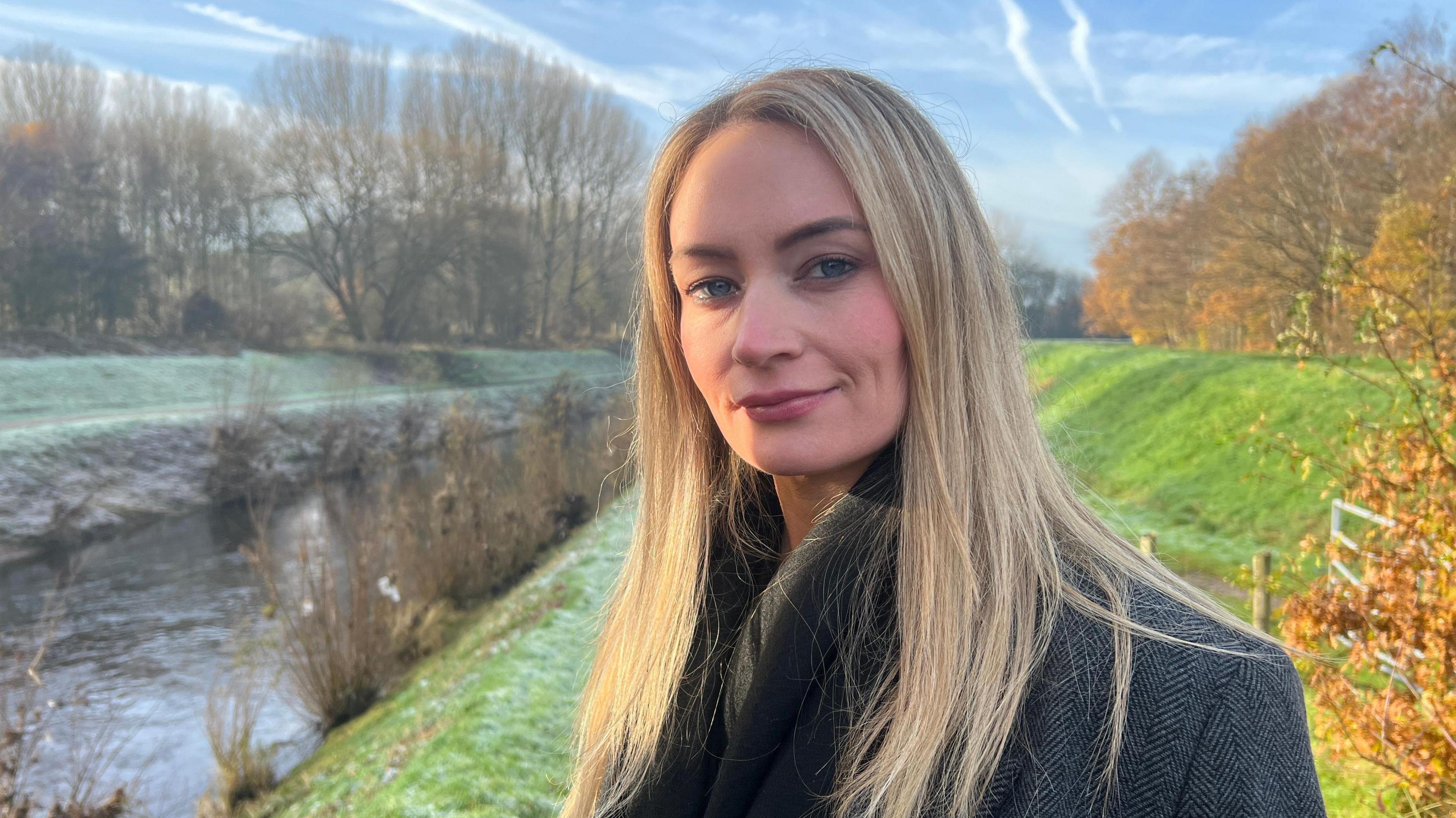 Detective Inspector Louise Edwards, who is a young woman with blonde hair, stands by a grass bank next to the River Mersey. 