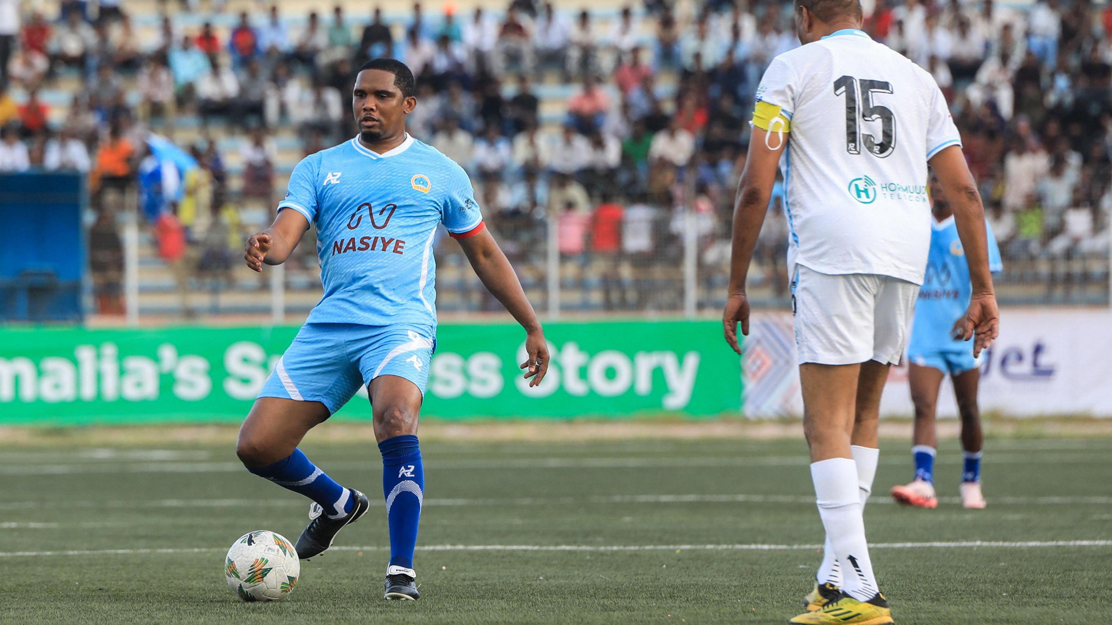 Samuel Eto'o wearing all light blue kit passes a ball during a football match while an opponent all in white stands nearby