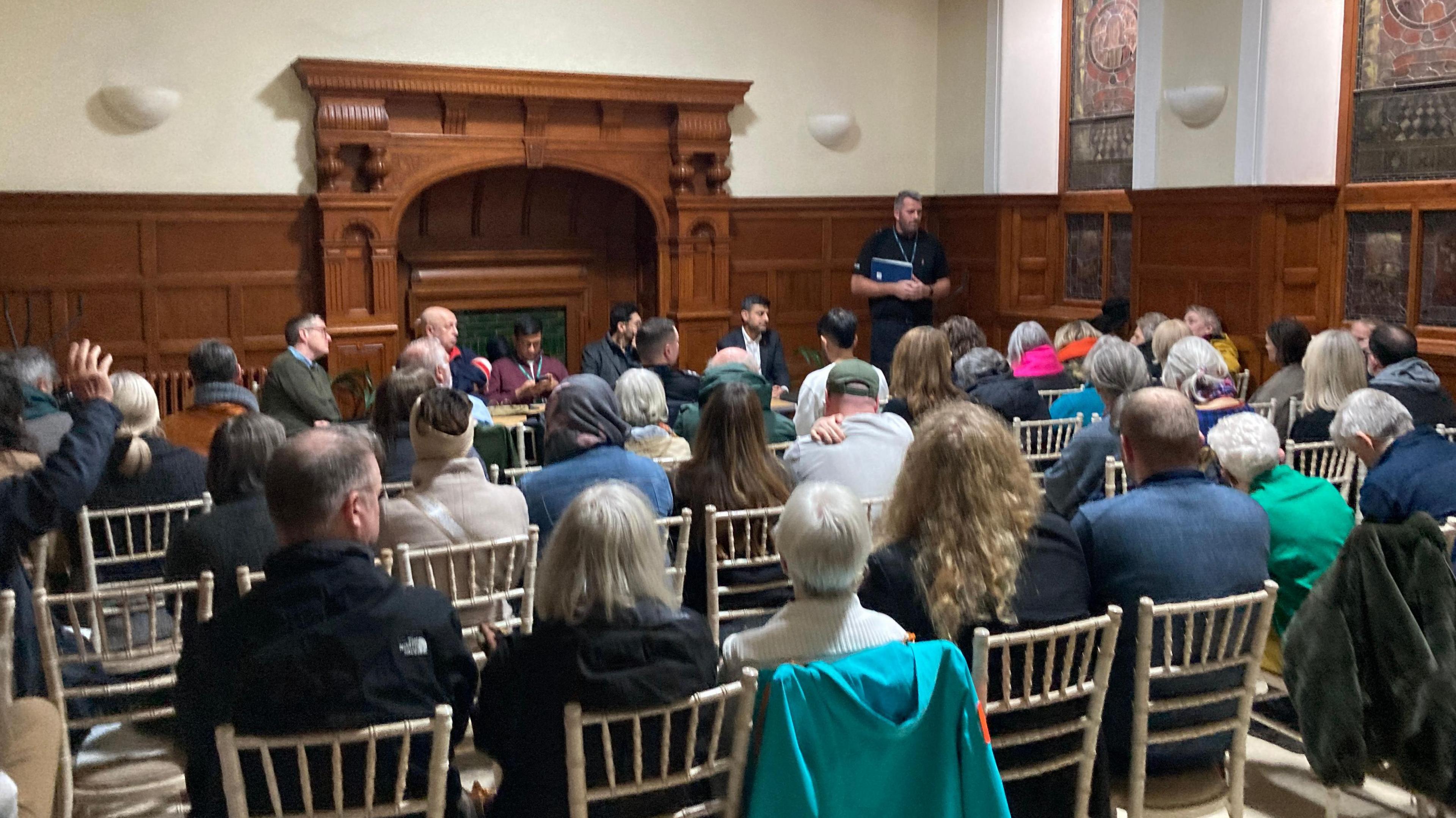 People sitting on white chairs listen to a police officer talking in a room with wood panelling and a grand fireplace.