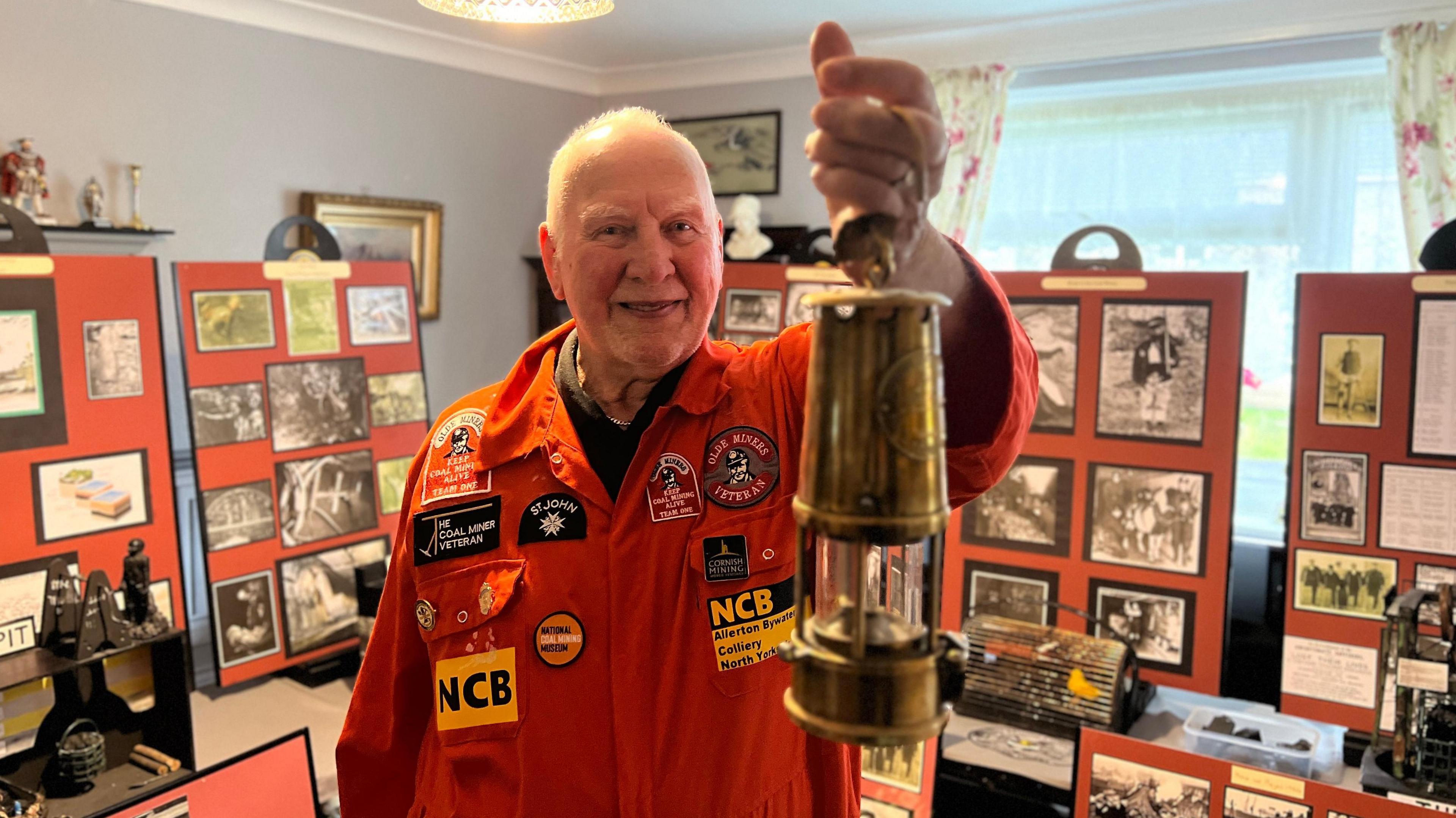 A man wearing orange mining overalls and with grey hair is holding a miner's lamp up in a room where behind him is a collection of mining pictures and artefacts.