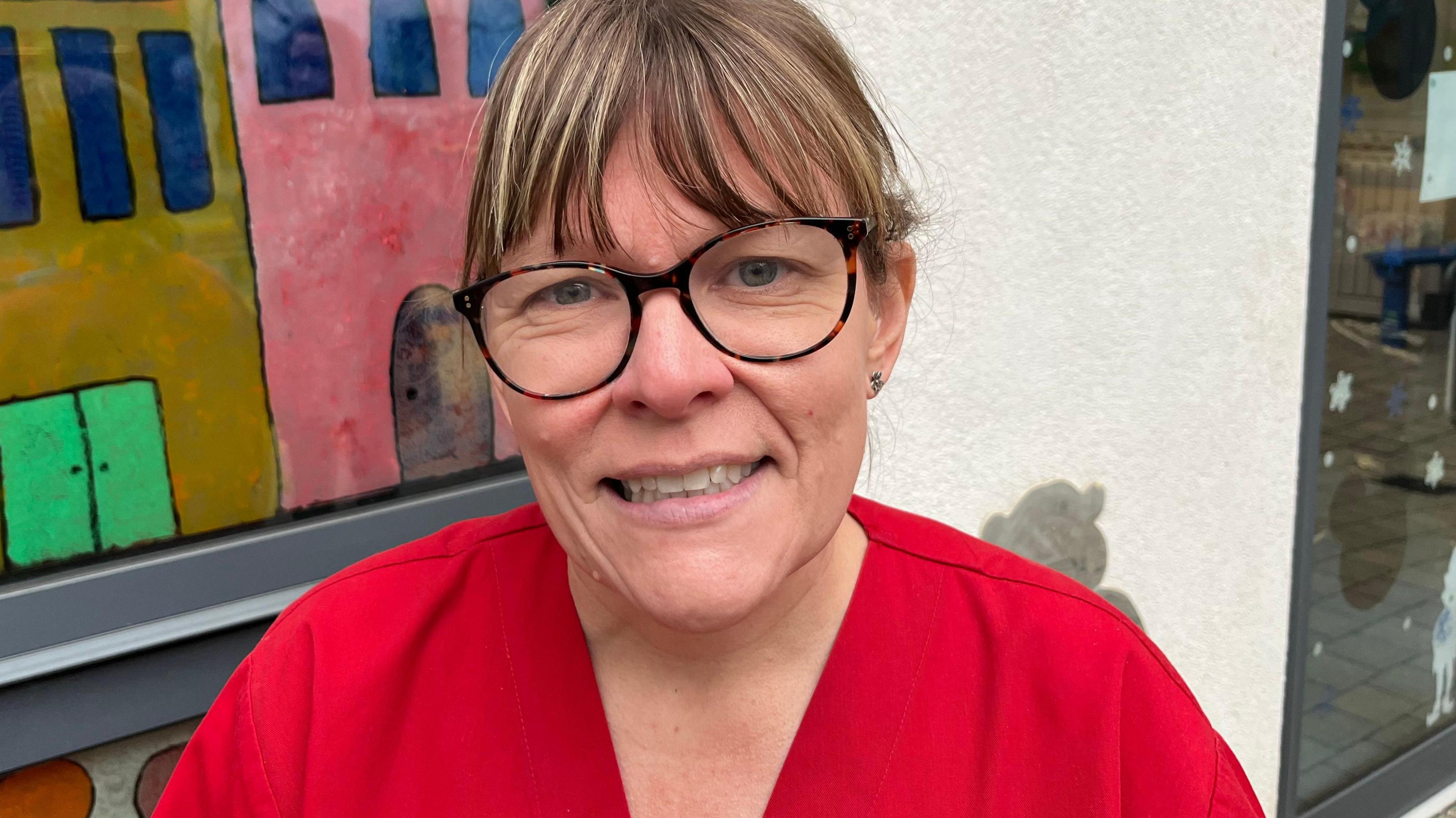 Zoe Wilkinson wears a red staff uniform, circle framed glasses and is smiling at the camera. She has dark blonde hair, which is tied up, and a fringe. 