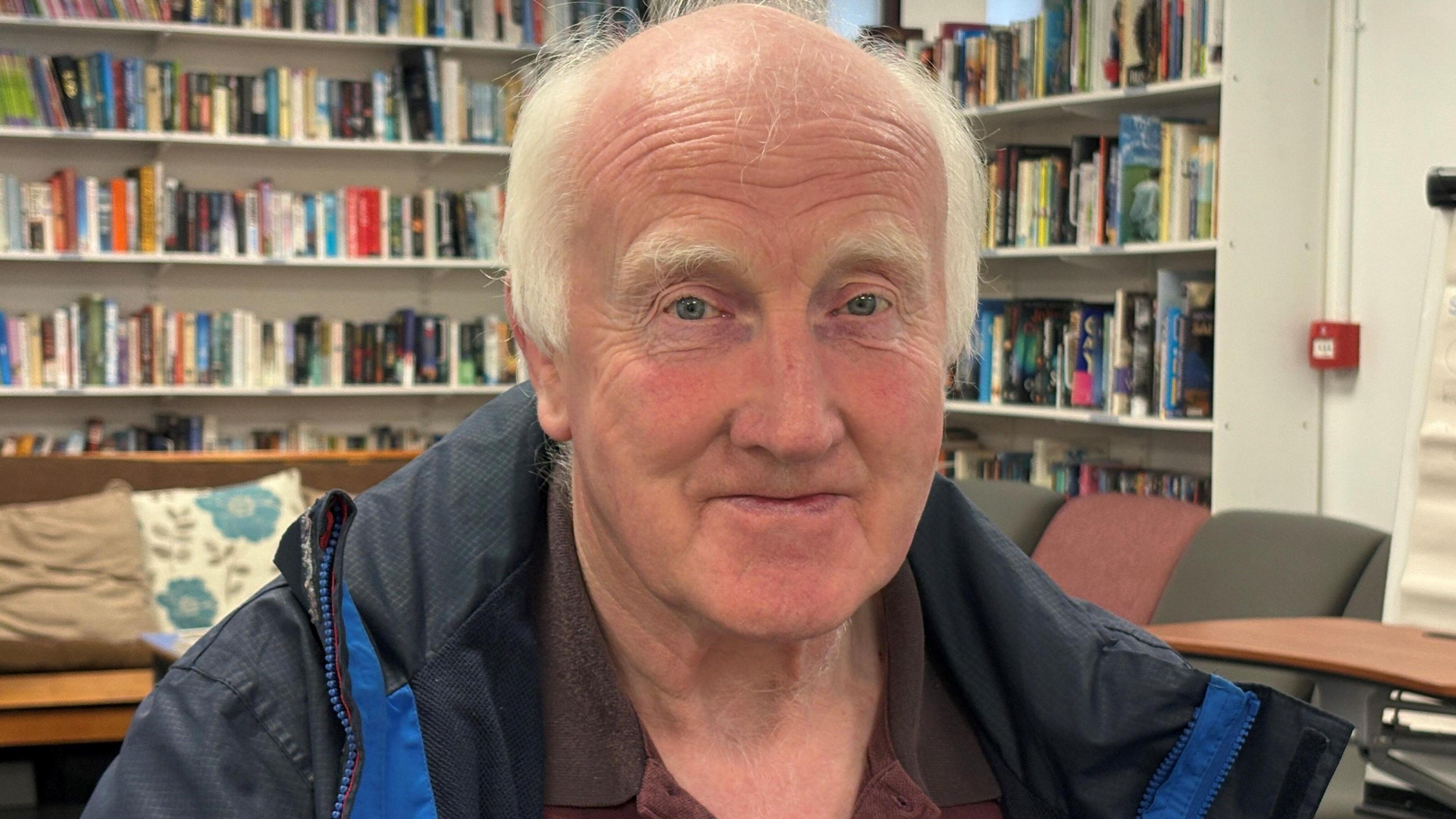 A man with white hair looks at the camera. He is wearing a raincoat. There are bookshelves behind him.