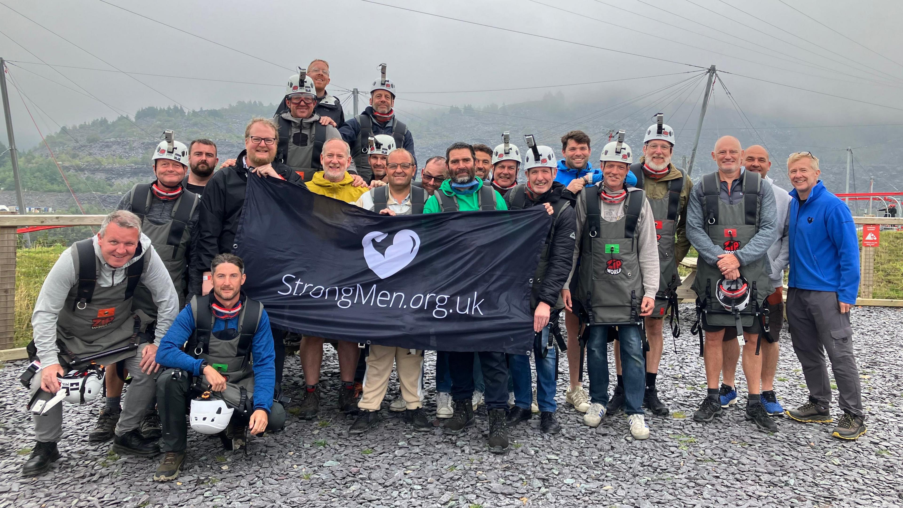 A group of men, some with helmets on, holding a large banner that says StrongMen.org.uk at an outdoor activity centre. 