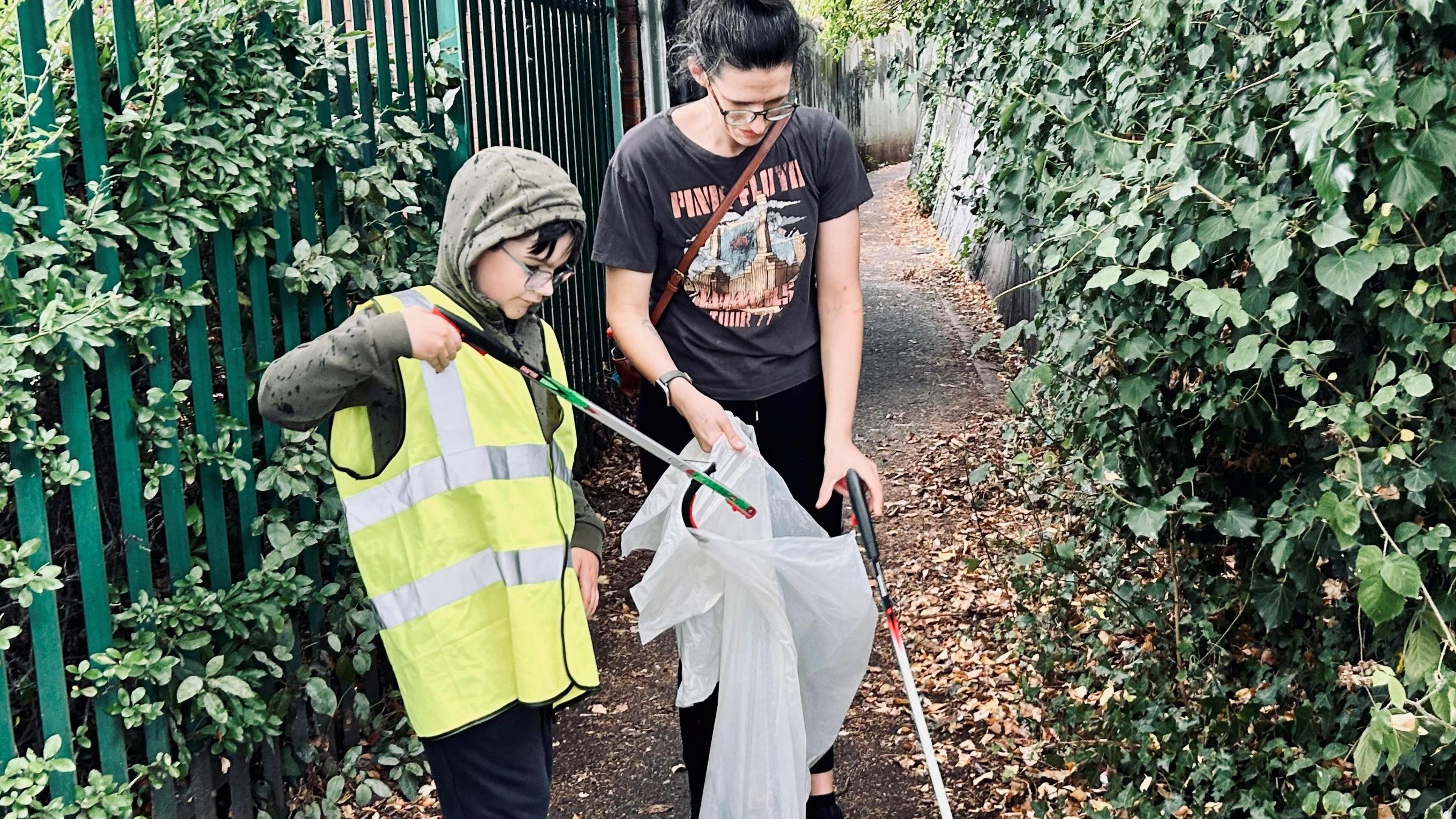 Eight-year-old Jackson - wearing glasses, a hi-vis gilet, green hoodie and black trousers - is pictured in an alleyway using a litter picker to put rubbish in a white bin bag. His mum is holding the bin bag open for him. She has black hair which is pulled back in a ponytail. There is a metal fence behind him with overgrowing leaves on both sides of the path. 