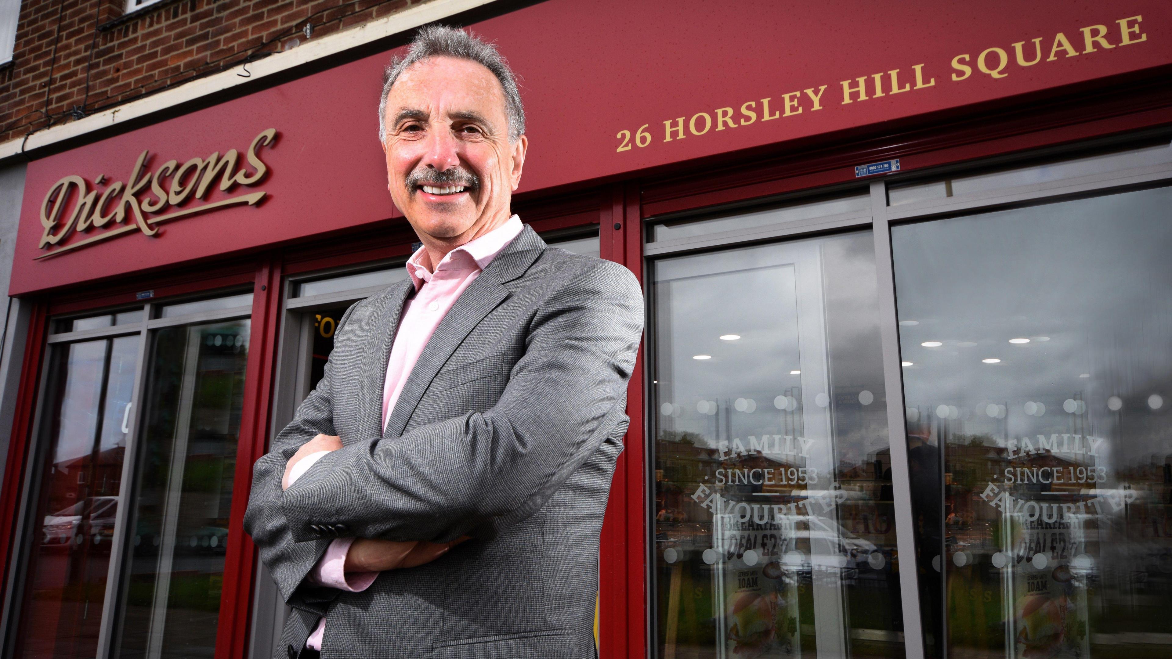 Michael Dickson, wearing a grey suit, stands outside a shop with a large Dicksons sign above his head