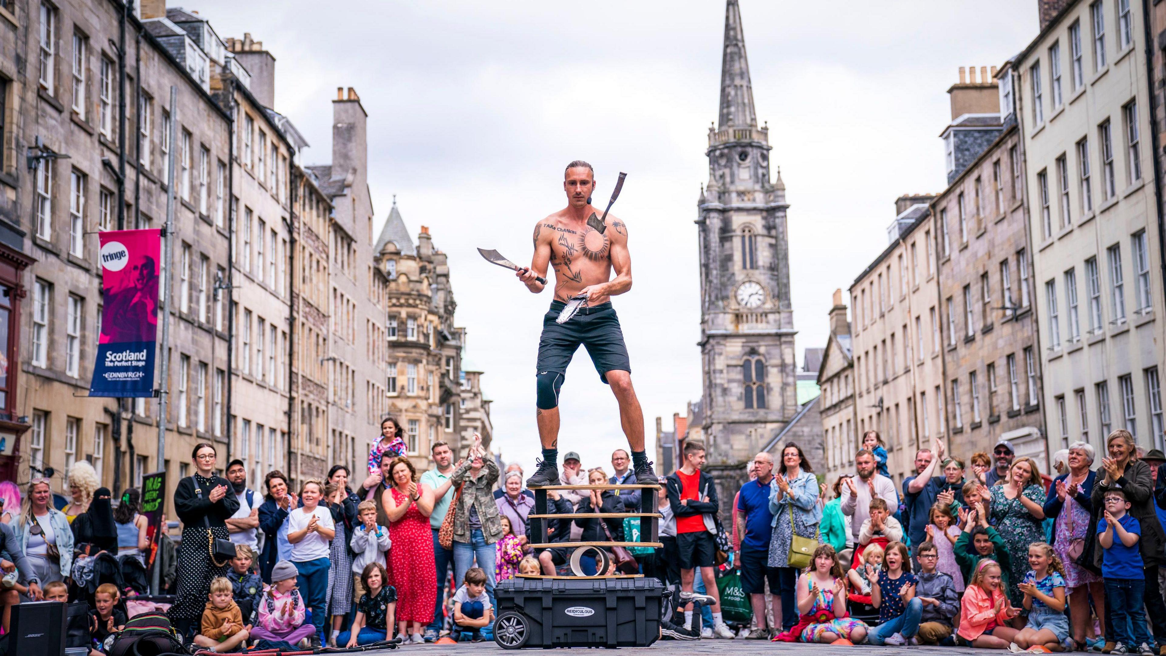A street artist juggles knives standing on a board balanced on a cylinder. Lots of people and children are standing or sitting watching. Monuments and historic buildings can be seen in the background.
