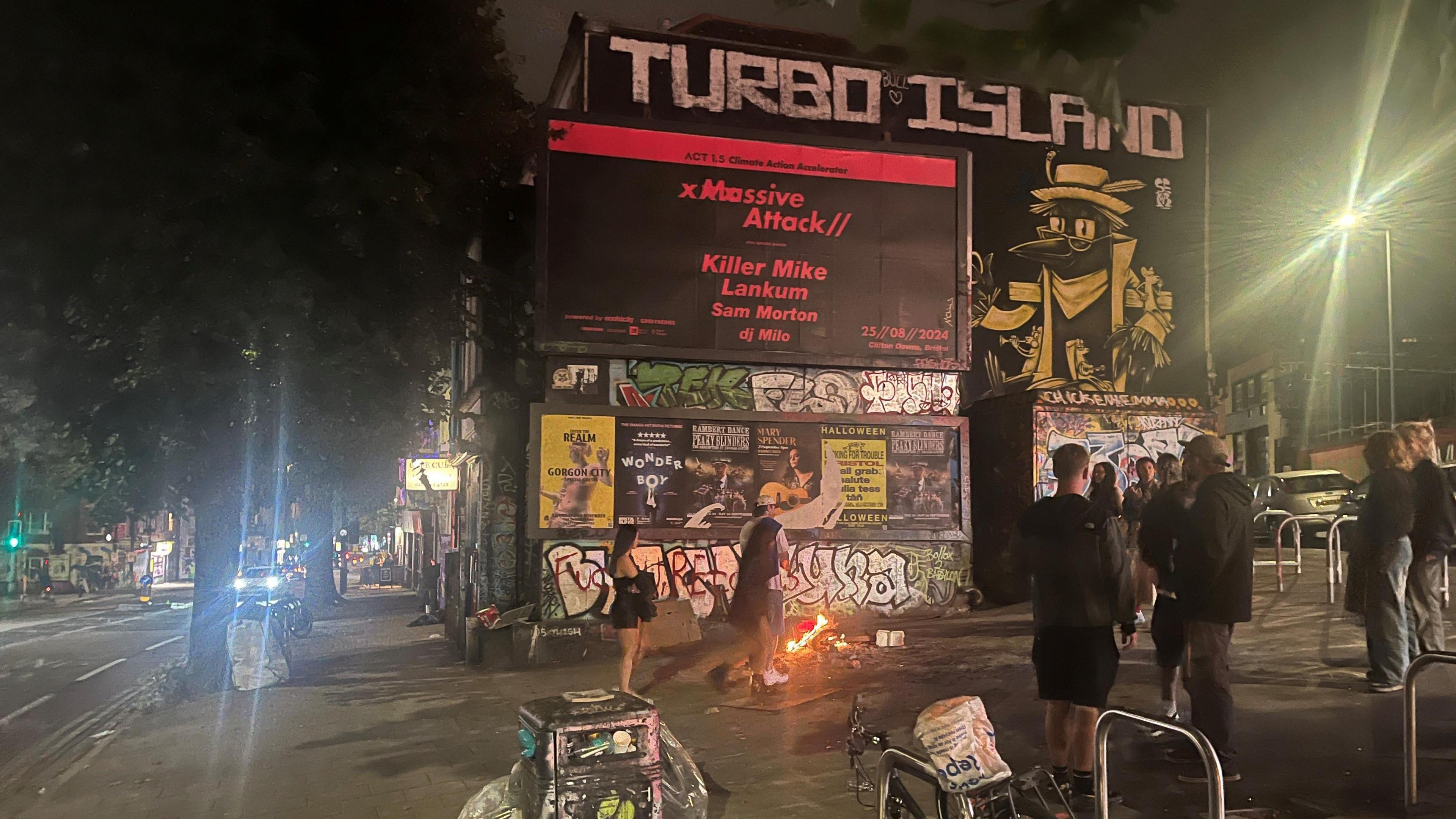 A piece of land located in a highstreet, with a graffitied sign on the wall which says 'TURBO ISLAND' and a billboard in front of it. There are people standing around a bonfire and some bike racks.