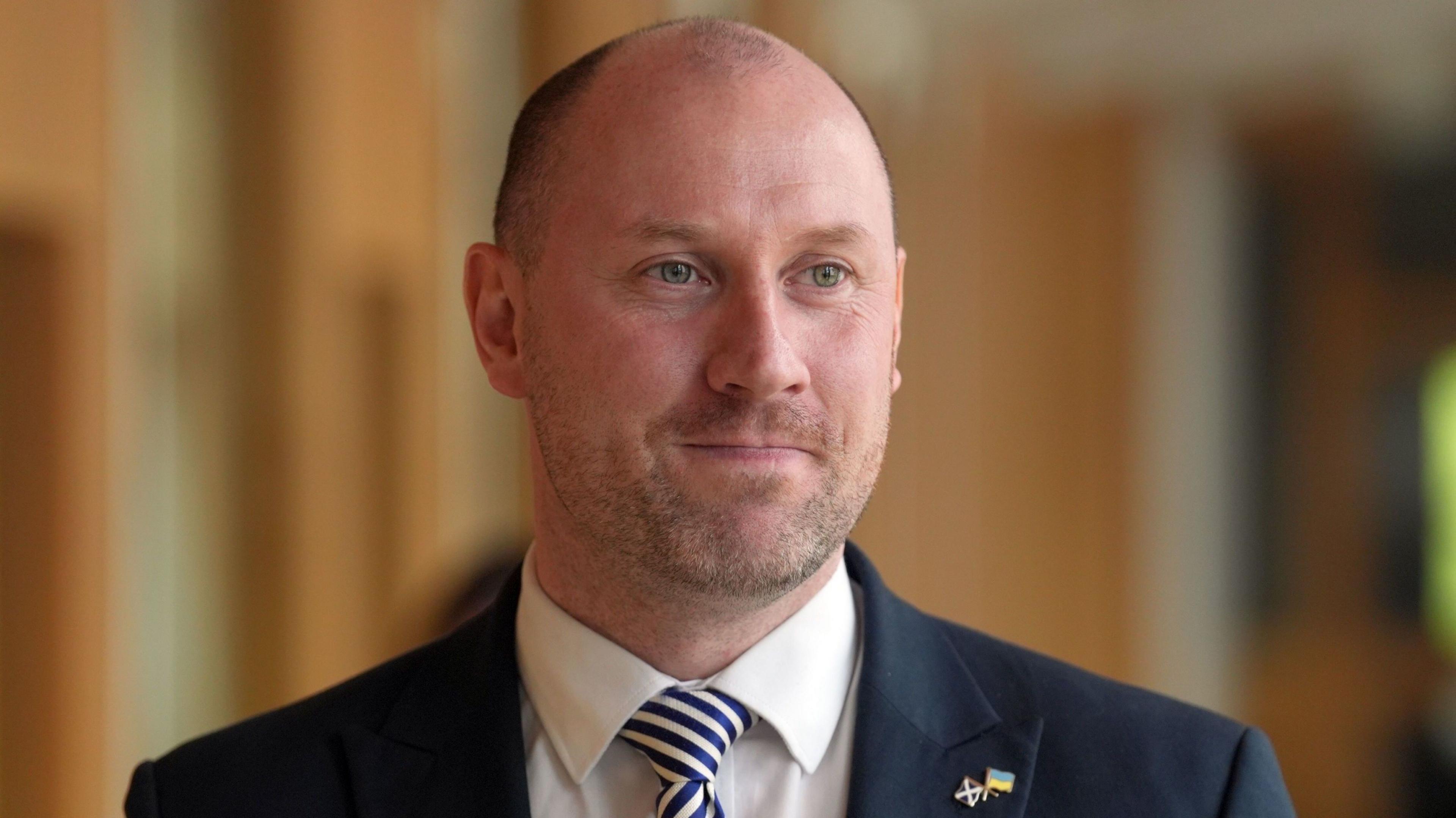 A bald man in a black suit, white shirt and blue stripy tie looks to his left in a close-up shot 