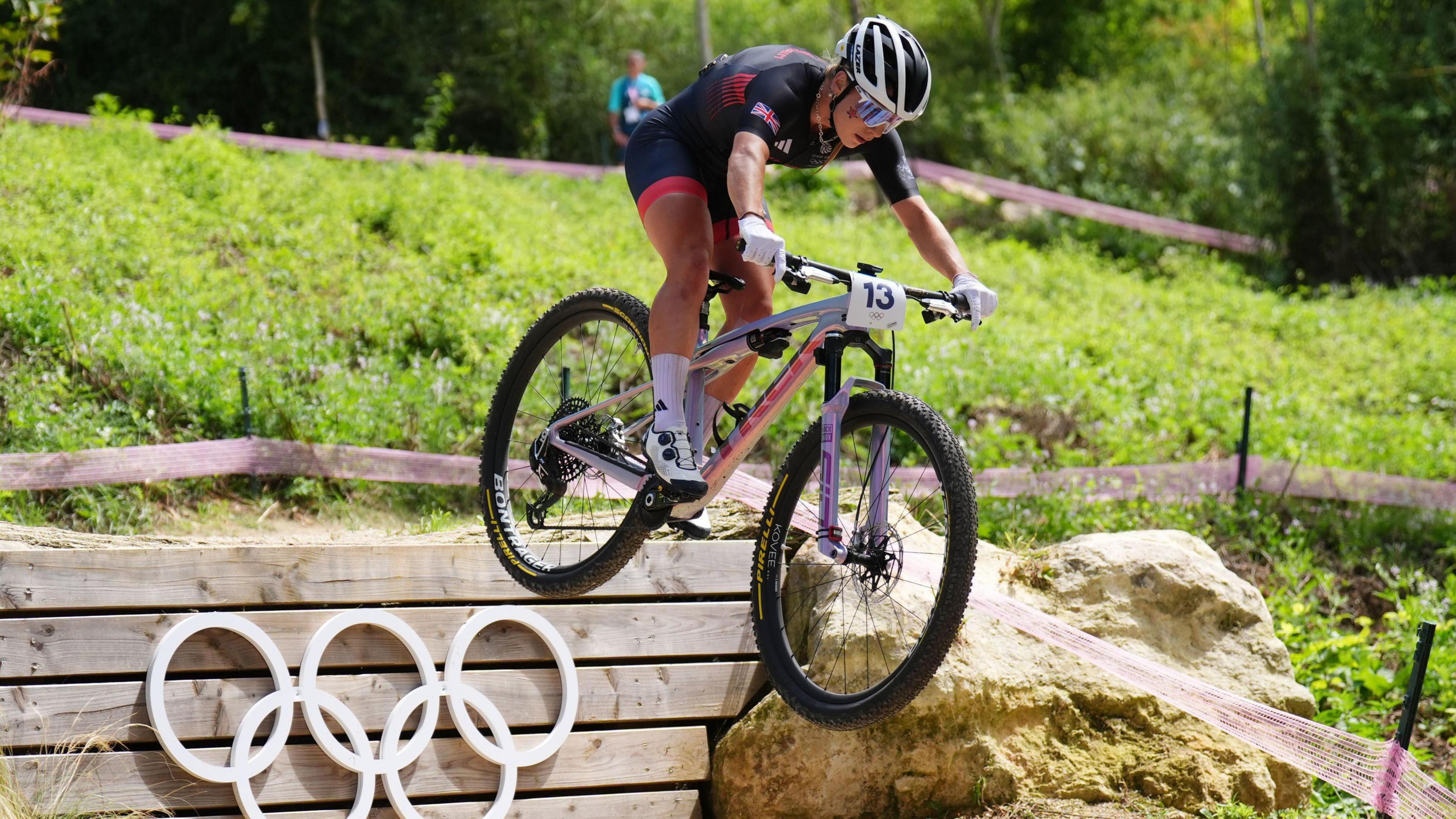 Evie Richards, on a white mountain bike is in mid air as she rides off a ramp. She is wearing a black and silver helmet, white gloves and a navy and red race suit. On the ramp are the five olympic rings.