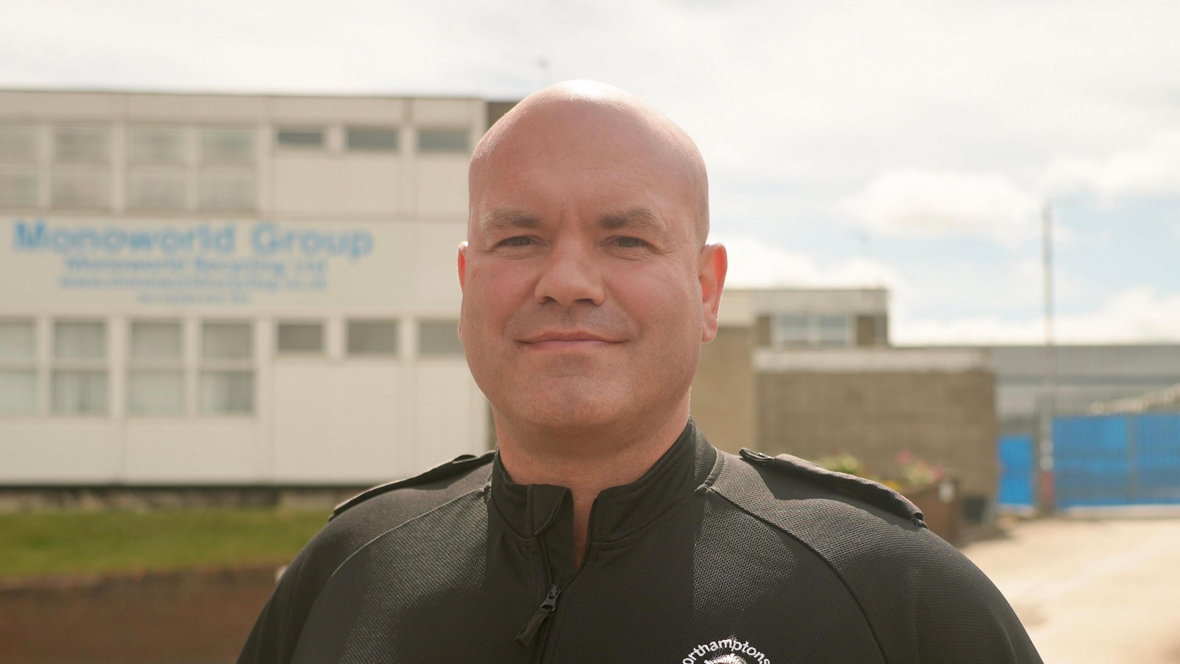 Daniel Retter with very short light-coloured hair smiling at the camera while wearing a black shirt with a fire service logo on it. There is a two-storey white building behind him with small windows and Monoworld Group in blue writing.