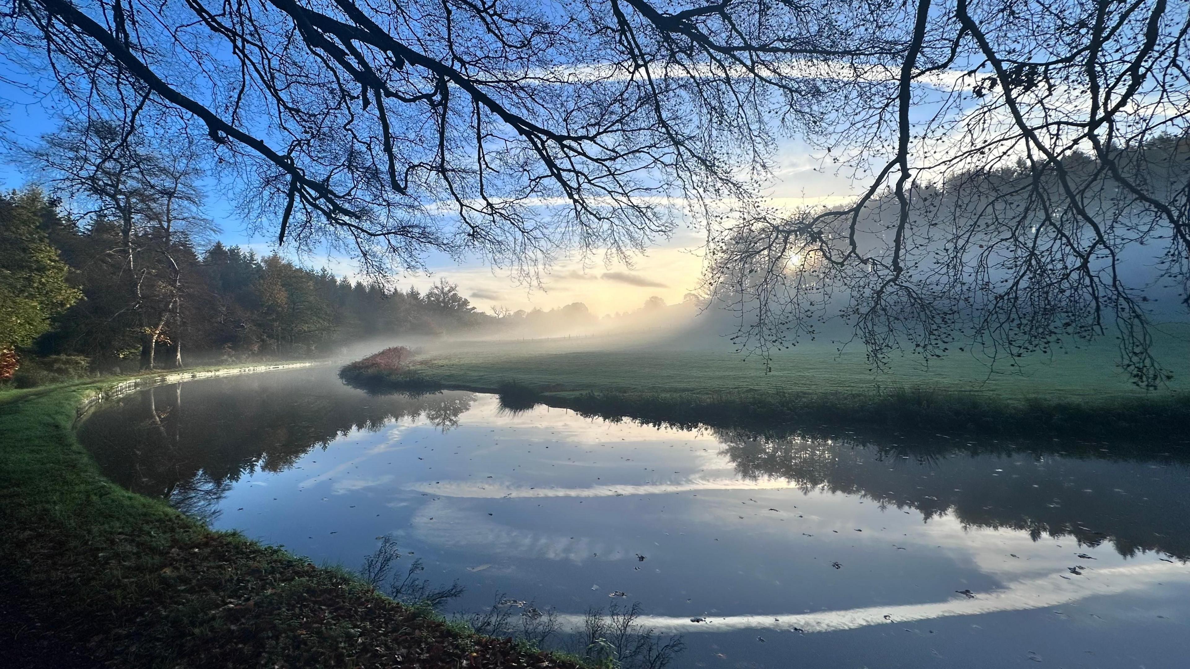 A wide canal looping around a field in Kinver. A mist hangs low over the water and the field on the far bank. Branches reach towards the water, above the photographer