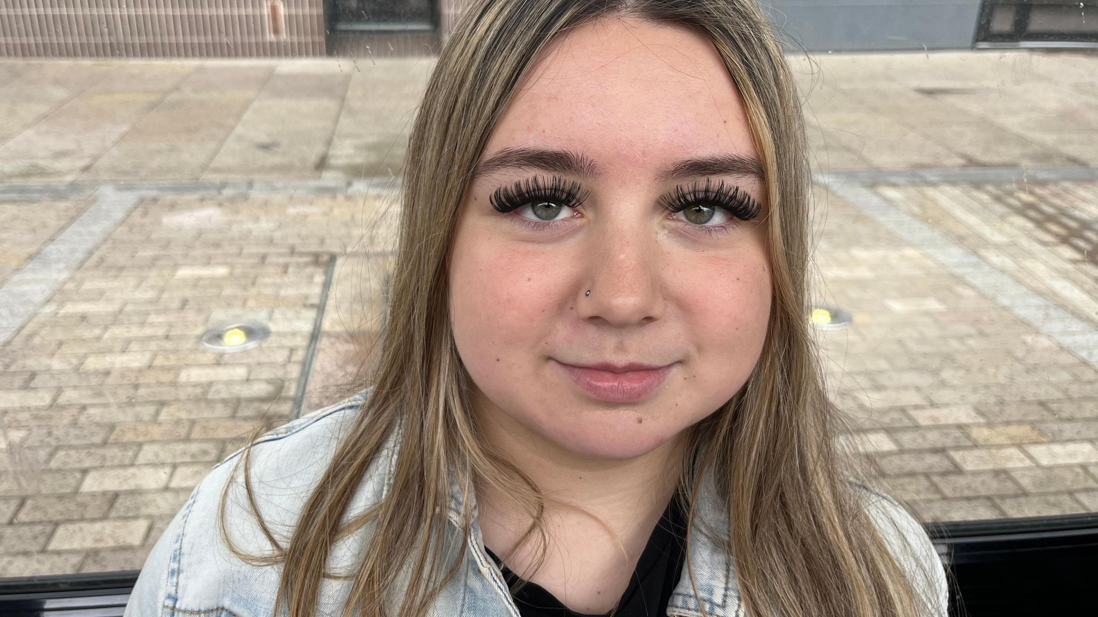 Paige Camplin who has long mid-brown hair and is wearing a light-coloured denim jacket over a black top. Behind her is block-paving.