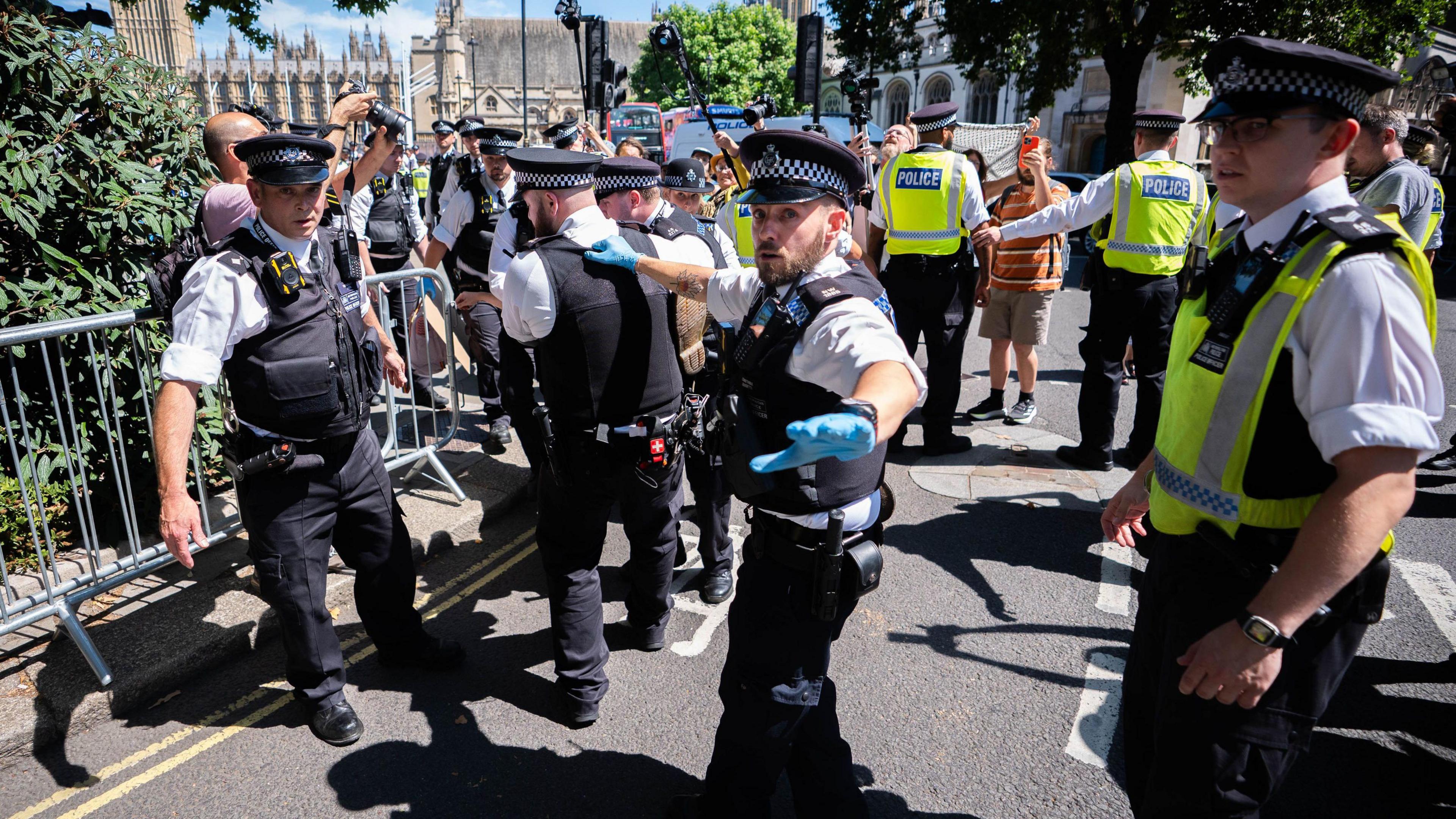 A number of police officers on a road with some members of the public taking photos and filming. The police appear to be carrying a person although only their foot can be seen in the image.
