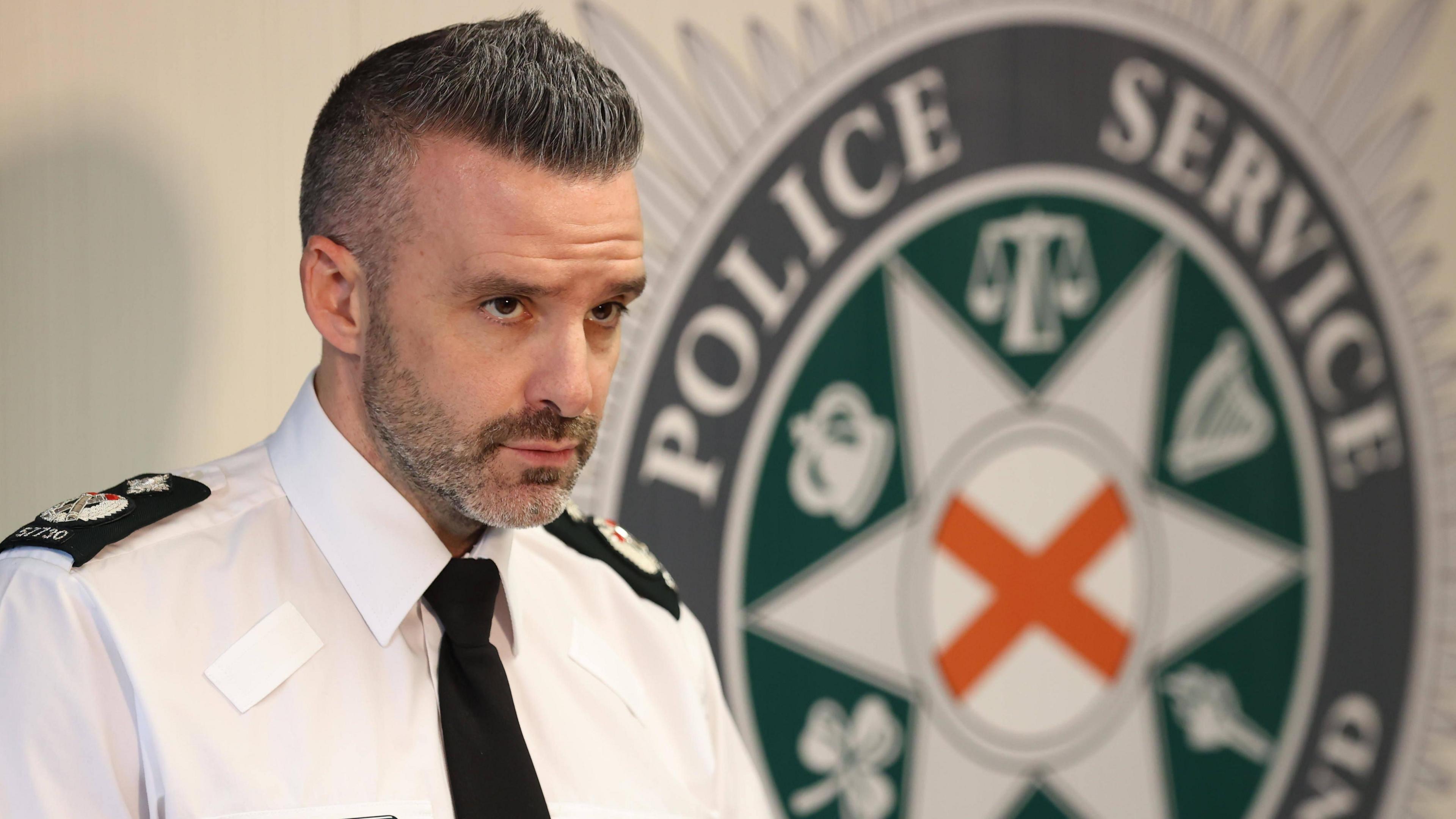Bobby Singleton stands at a press conference beside a PSNI crest. He is wearing a police uniform - a white shirt with a dark tie and shoulder lapels. He has short, dark, greying hair and heavy stubble. 