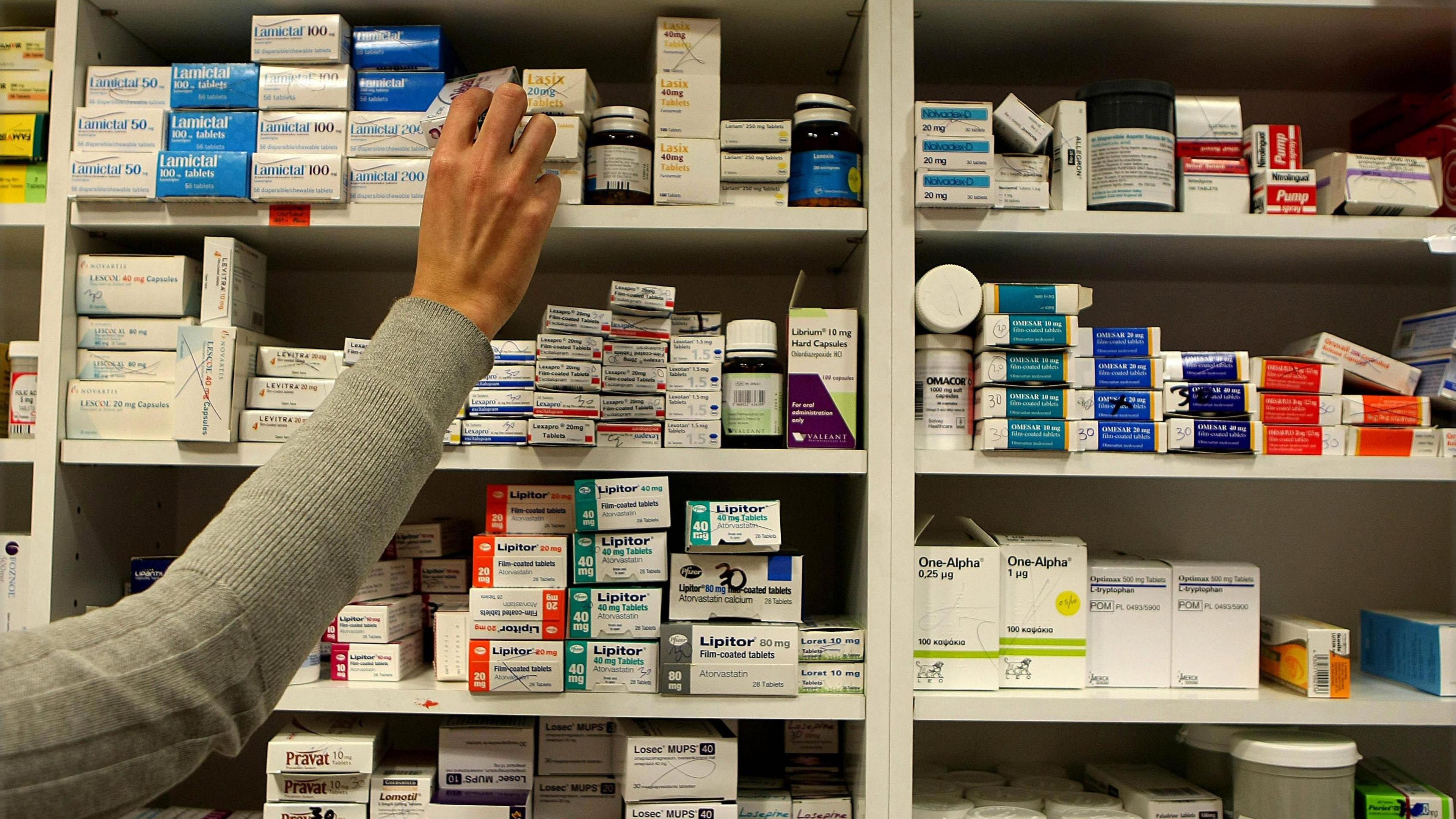 A woman's arm is stretched up to shelves piled high with different medications and she is pulling a small box of one of the higher shelves.