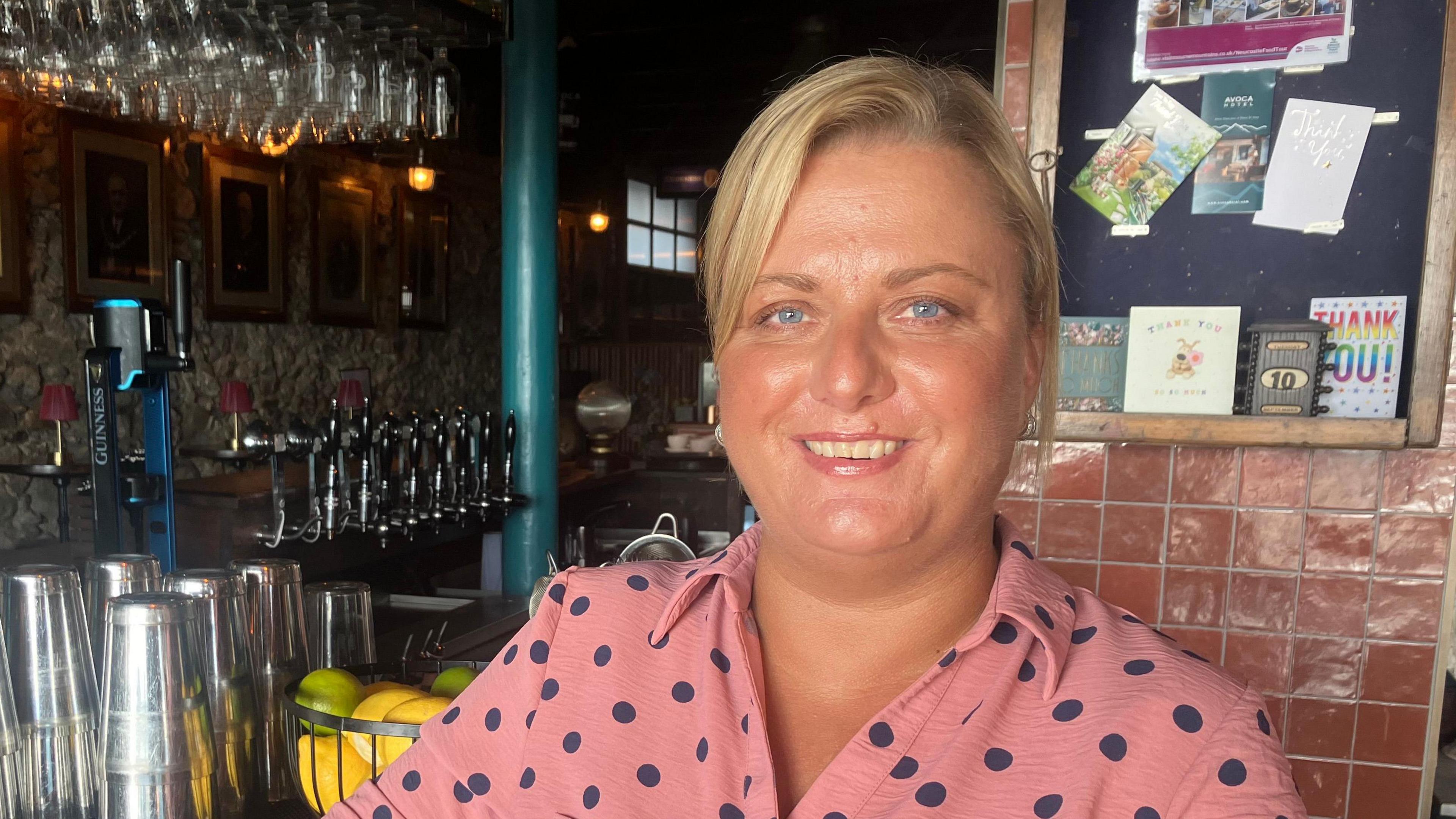 Nicola Stevenson is wearing a pink polkadot linen looking shirt. 

She has blonde hair tied up in a pony tail. Her hair is in a side parting and she has blue eyes. She is smiling at  the camera. 

She is standing in front of a bar stacked with wine glasses and draught beer. Beside the bar there is a fruit bowl and plastic cups. 