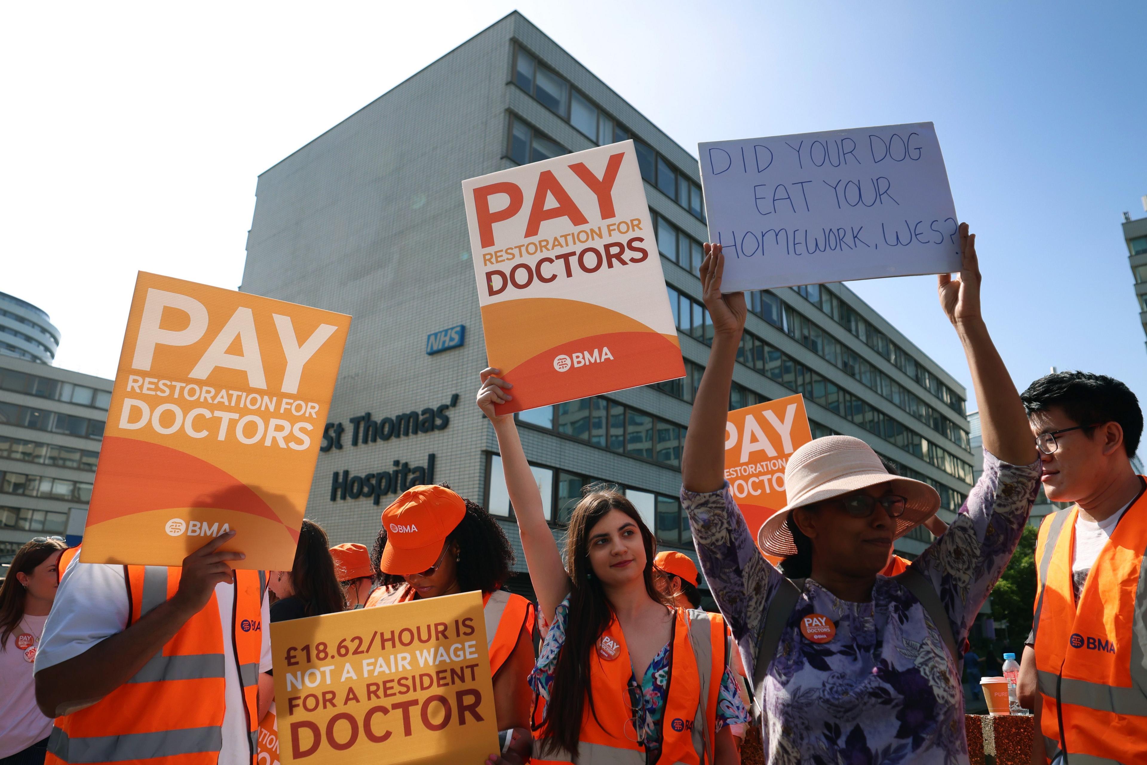 A group of resident doctors on strike hold orange placards calling for pay restoration outside St Thomas's hospital in London, which is visible behind them.