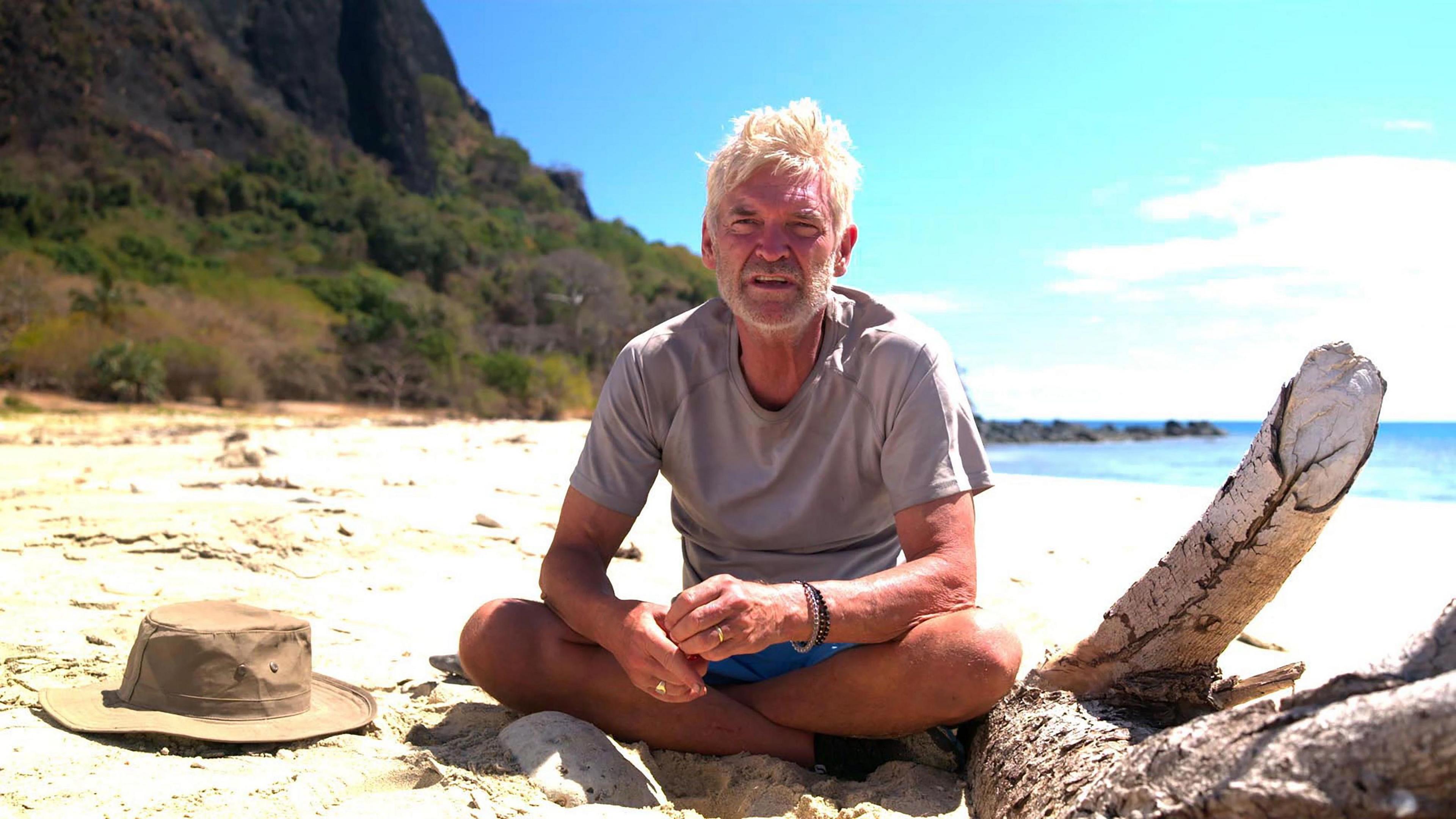 Phillip Schofield sitting on a beach in Cast Away looking pensive in shorts and T-shirt 