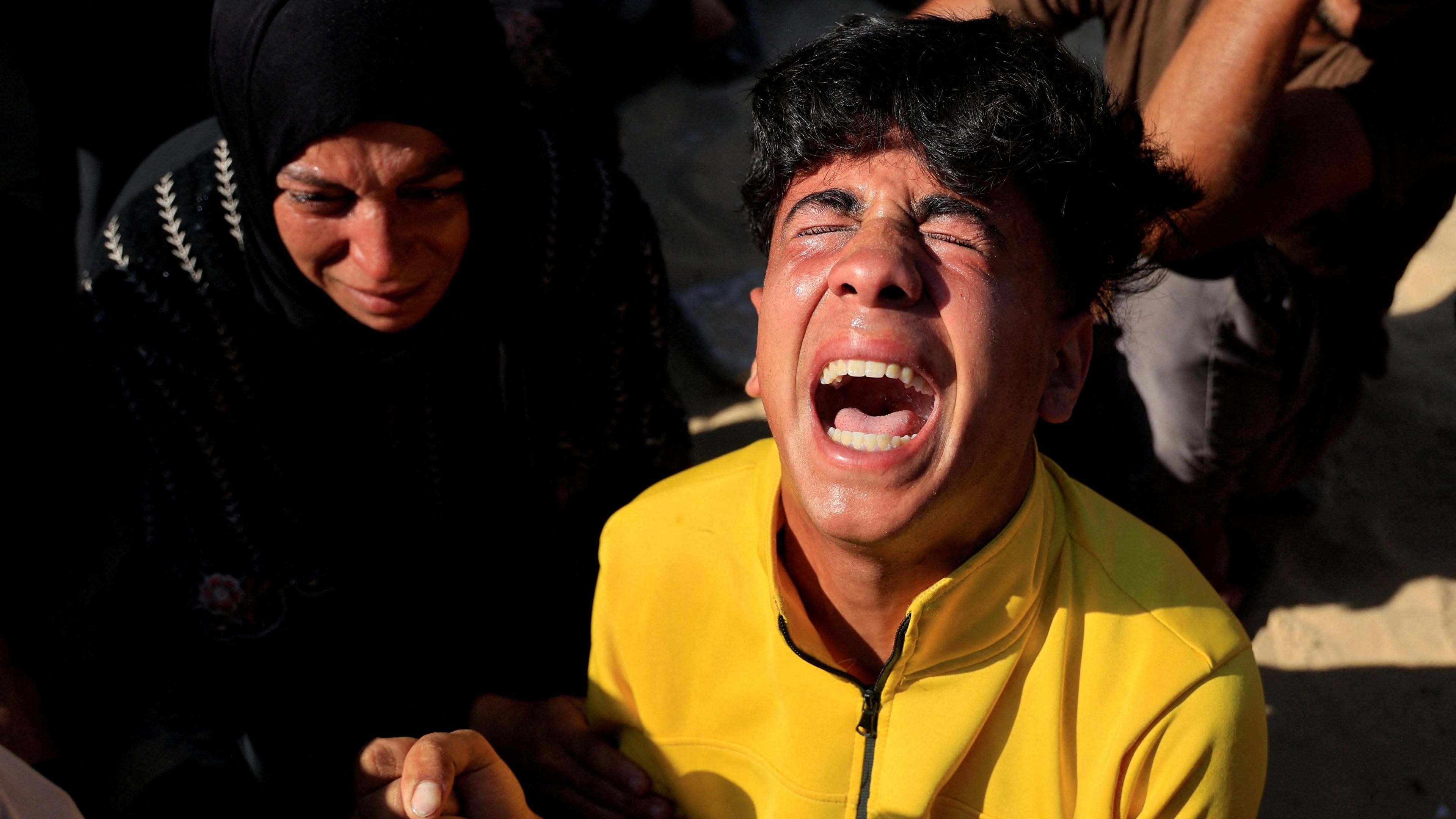 A young man wearing a yellow jumper cries, his eyes clenched closed and his mouth open, as a woman beside him in a black abaya and hijab looks down.