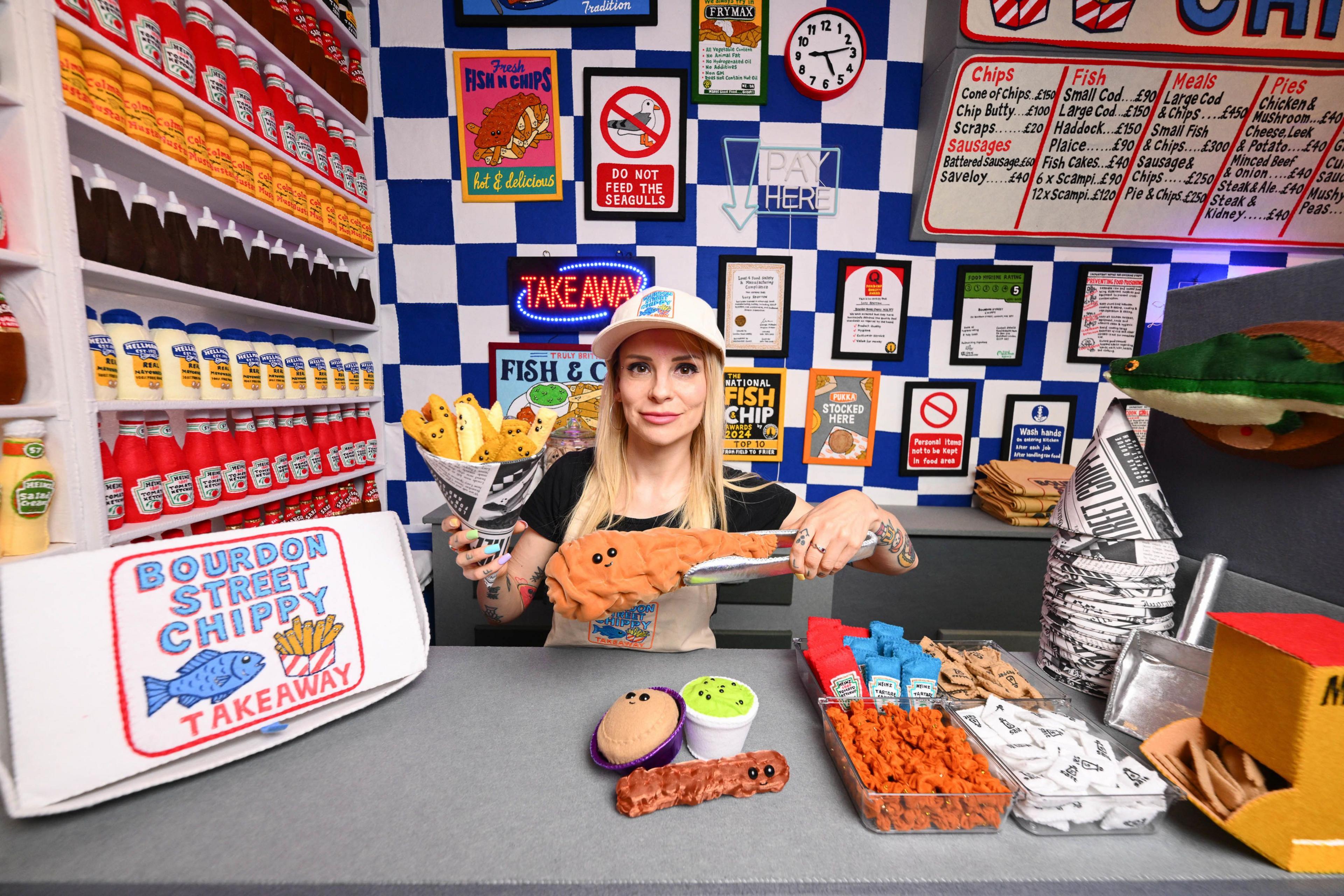 Artist Lucy Sparrow stands behind the counter of her fish and chip shop, holding a large felt-made piece of fish.