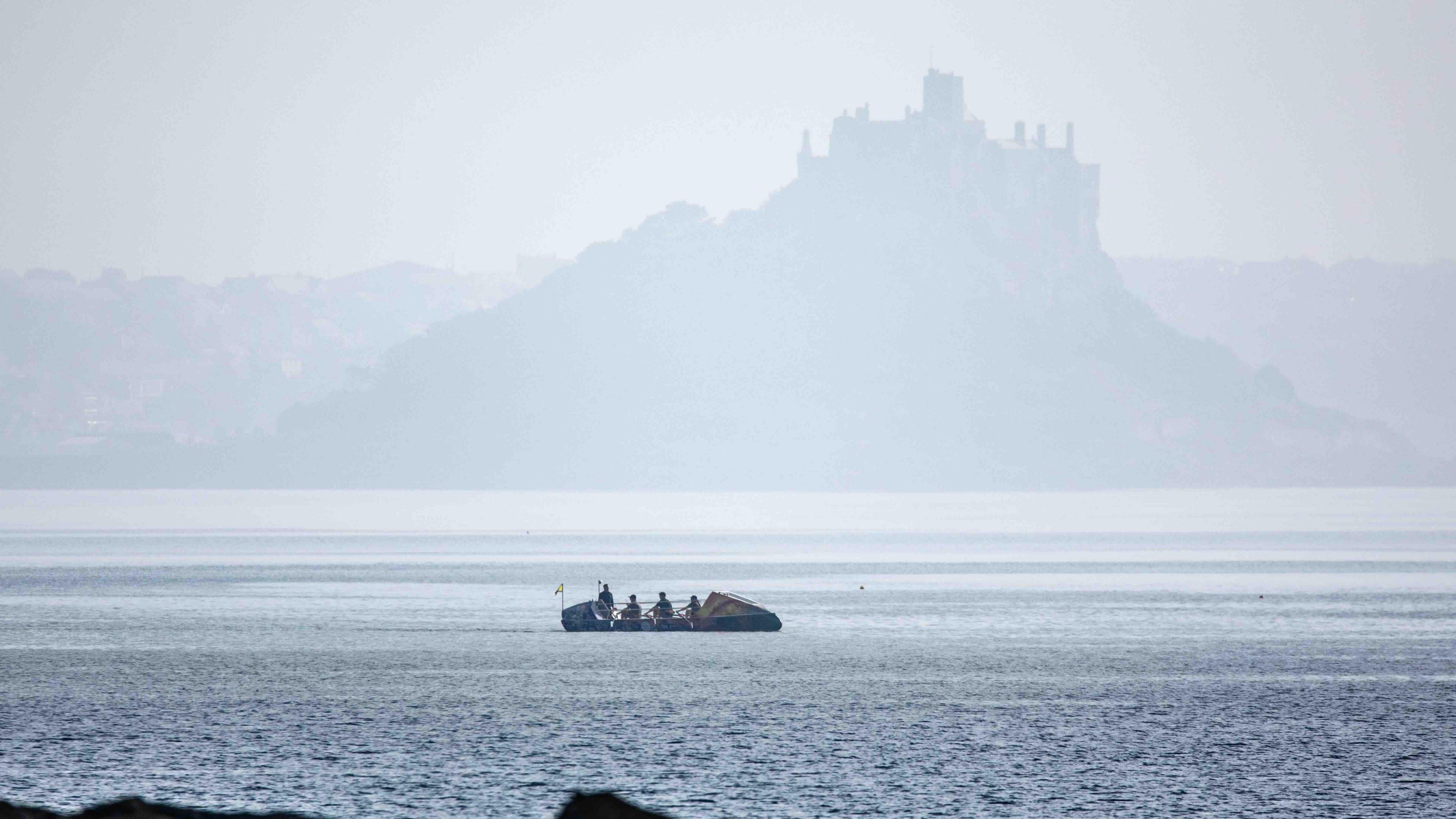 An ocean rowing boat with four crew passes a large island and castle.