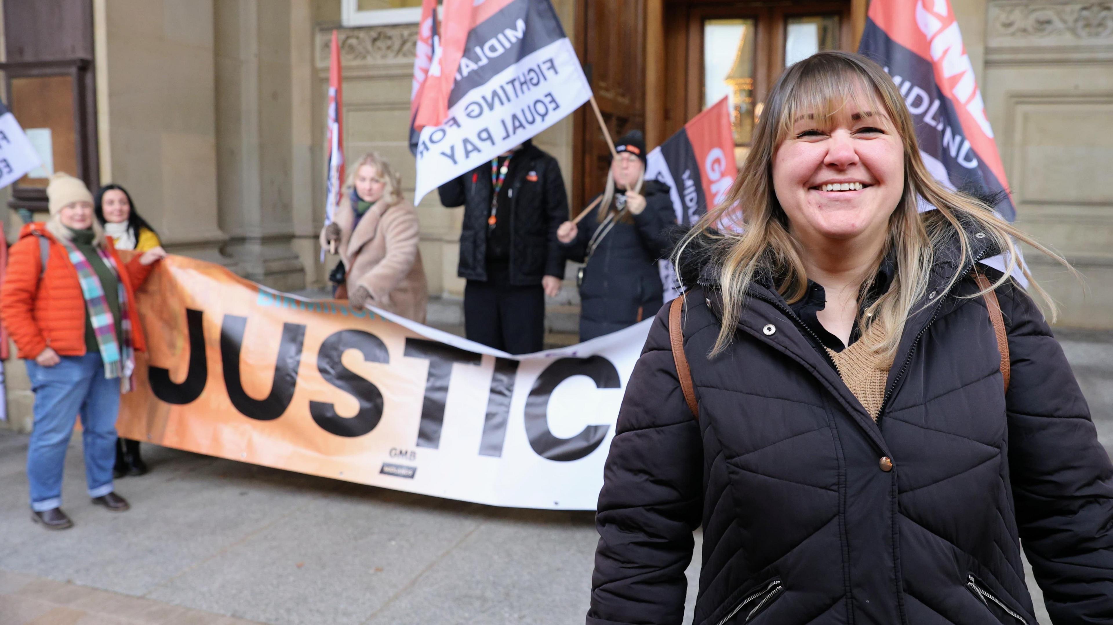 A woman in a black puffa coat standing outside a sandstone building with ornate carving on some bricks and stone pillars. Behind her stand four people holding a banner and flags saying "Justice and Fighting for Equal Pay"