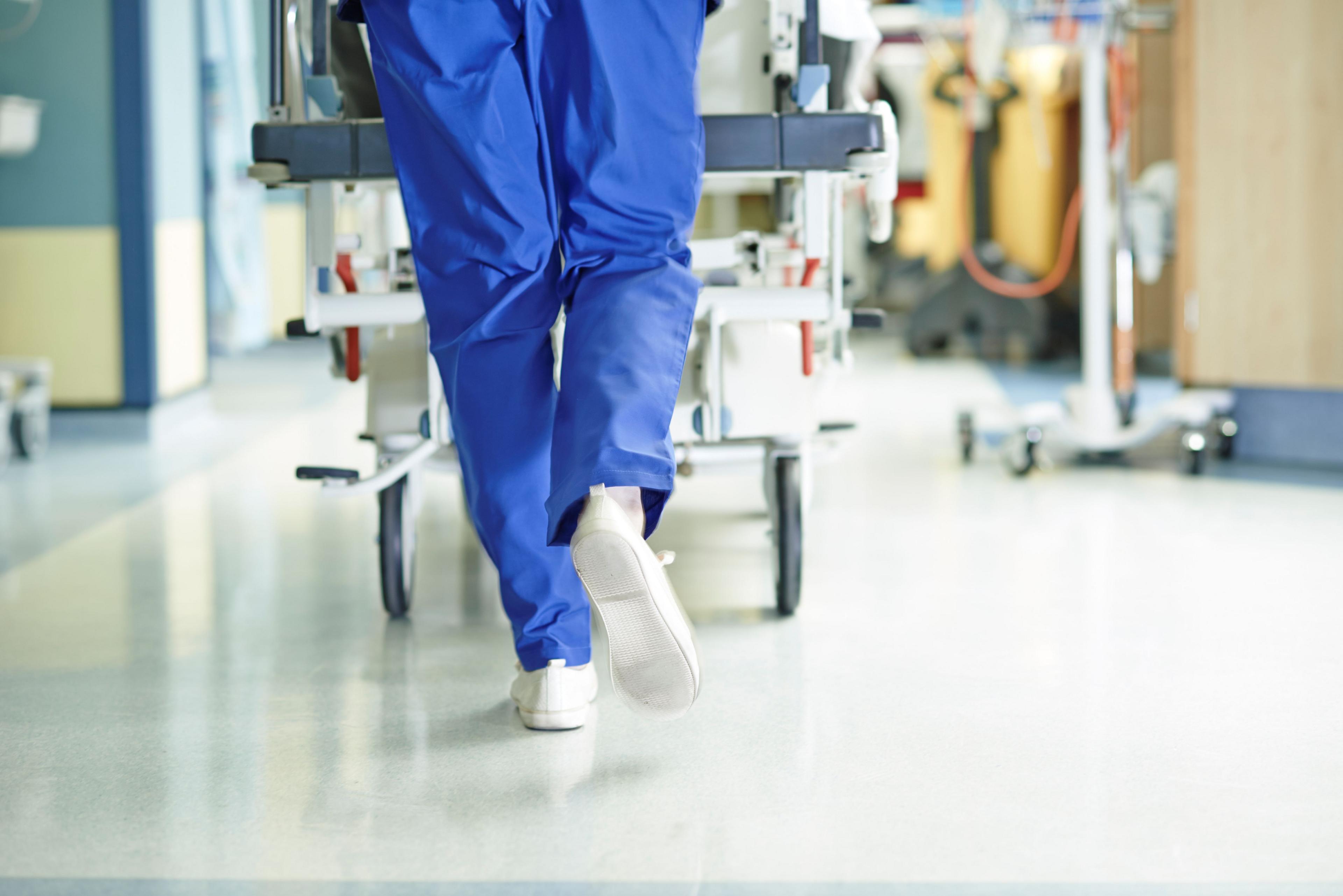 A medical professional pushes a trolley down a corridor. Their white trainers and blue trousers are visible. They are walking along a polished floor.