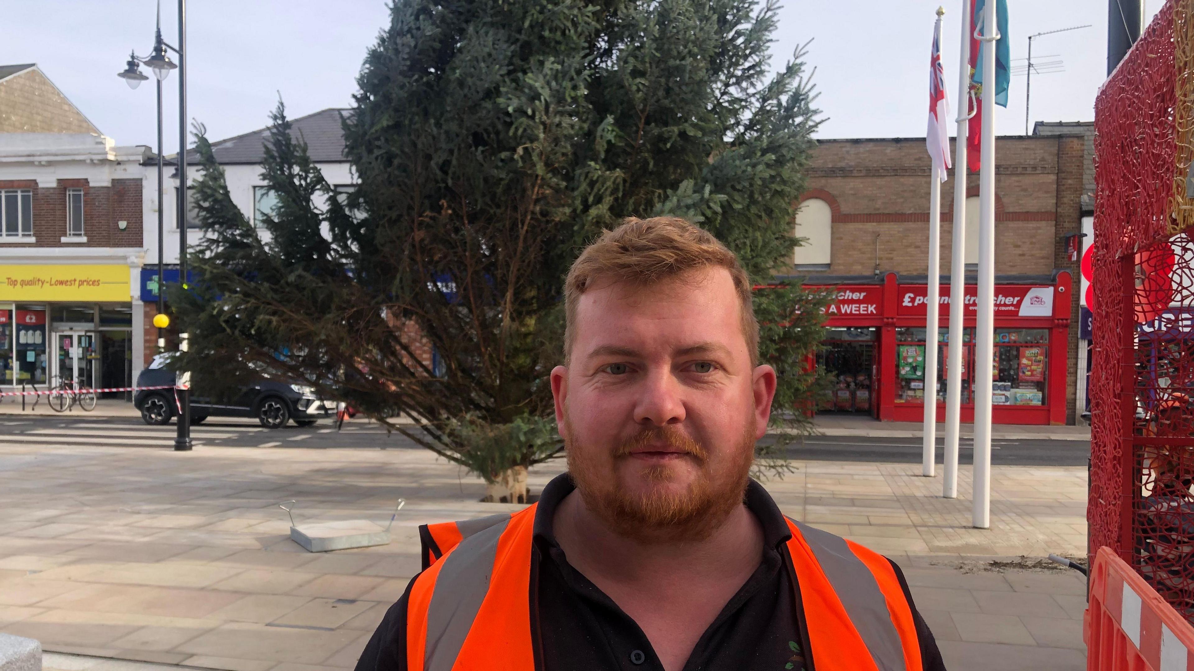 Daniel Fortuna has red hair and a beard with a moustache. He has a black T-shirt and a hi-vis orange tabard. Behind him is the bottom section of the Christmas tree, which is leaning slightly to the right. There are flagpoles and part of the town's war memorial on the right, with shops in the background