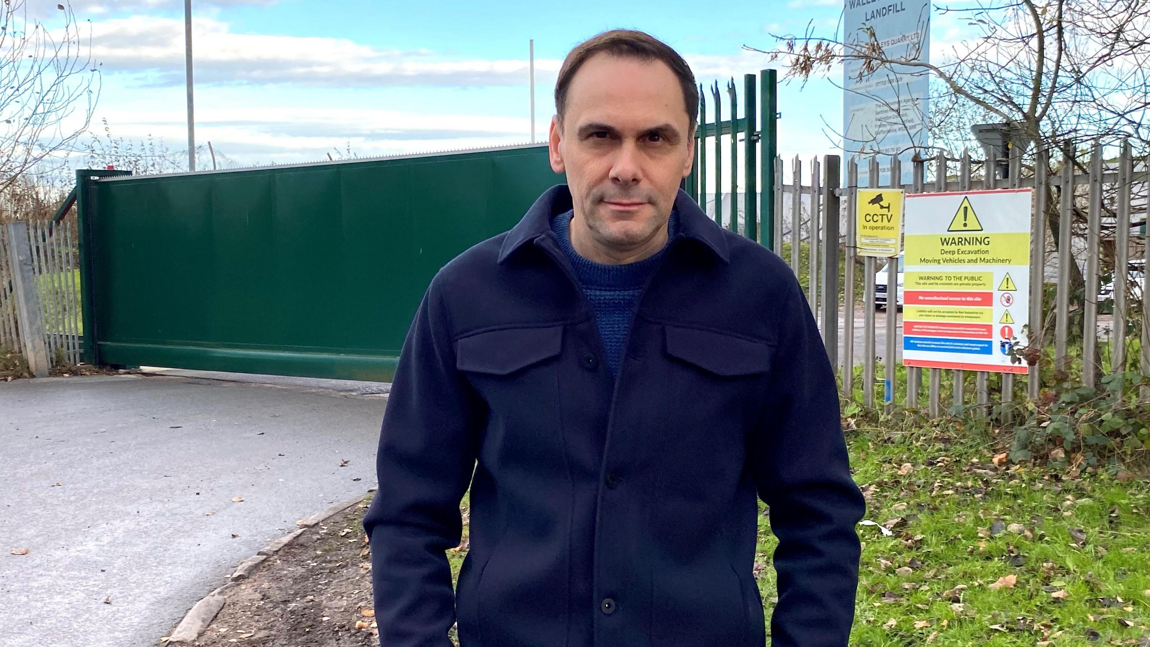 Simon Tagg, a man wearing a navy blue coat, is standing outside a large set of green gates at the entrance to a landfill site. There is signage on fencing to the side of the gates which indicates the site is hazardous.