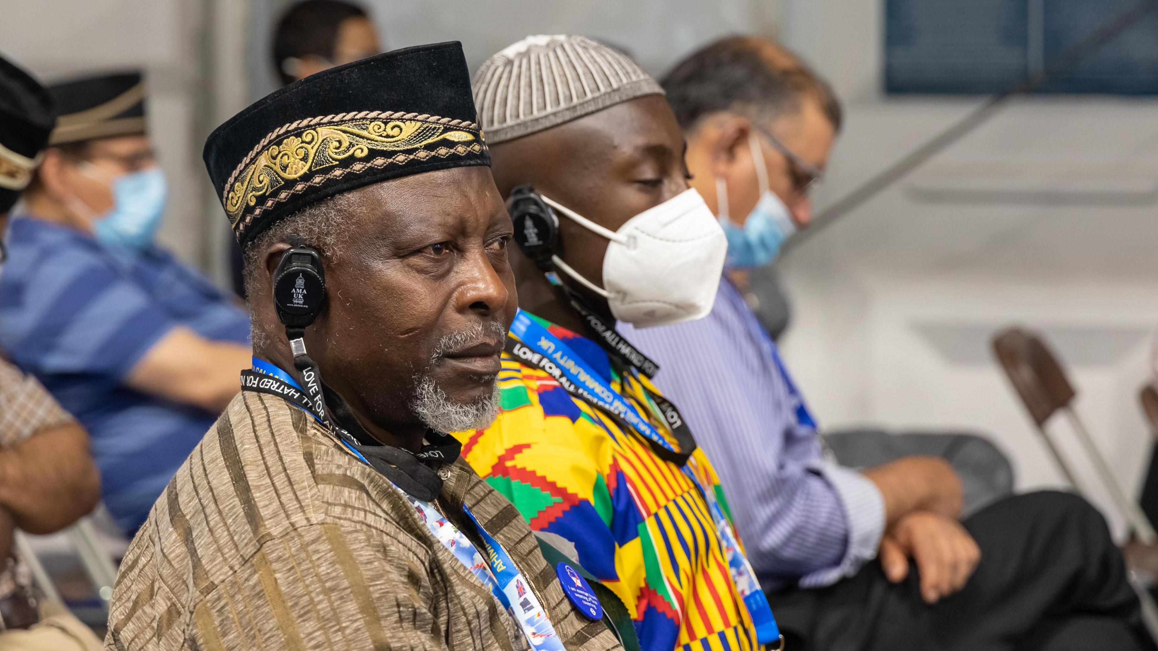 Men from different ethnic backgrounds are seated next to each other at an indoor seminar. Two of them wear headphones.