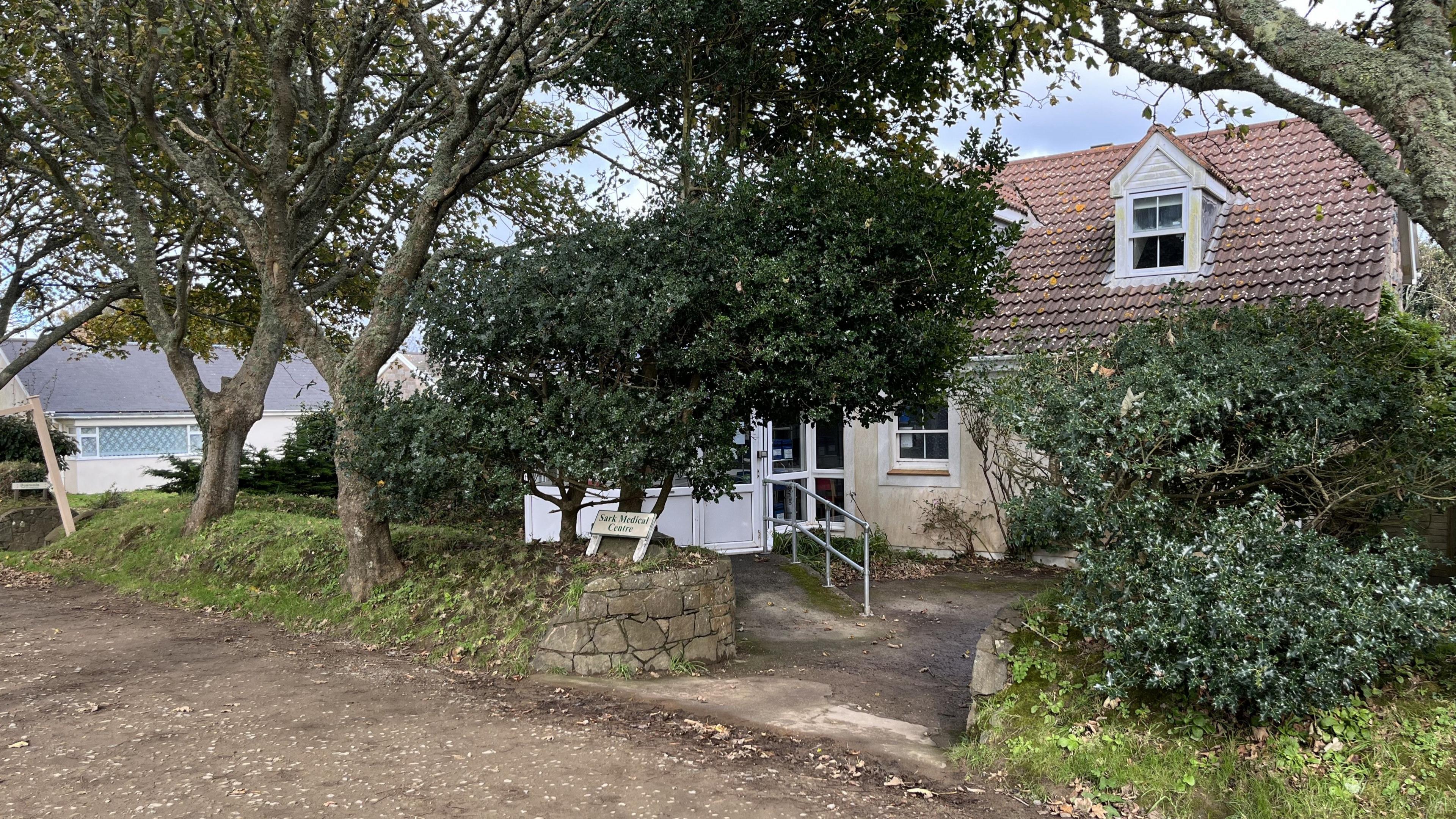 A small, two-storey, cream building with a reddish-brown tiled roof sits next to a dirt path, partly obscured by trees and bushes. A UPVC porch has a ramp and railing outside. A small white wooden sign in the ground below a holly tree. The sign is off white with green writing reading Sark Medical Centre.