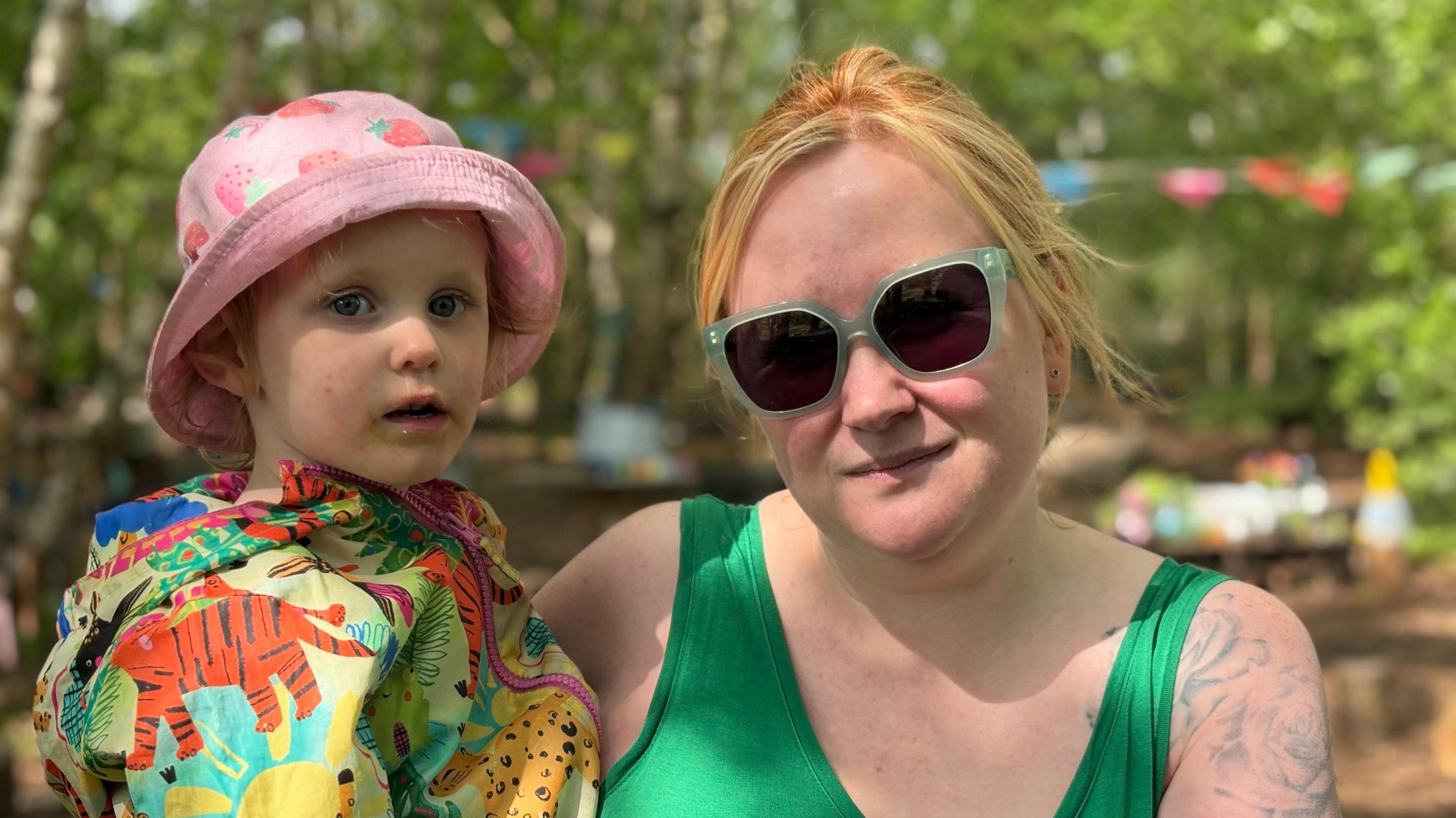 A woman in her early thirties with red hair wearing green sunglasses holding her infant daughter. They are in a woodland area with green leaves on trees visible behind them.