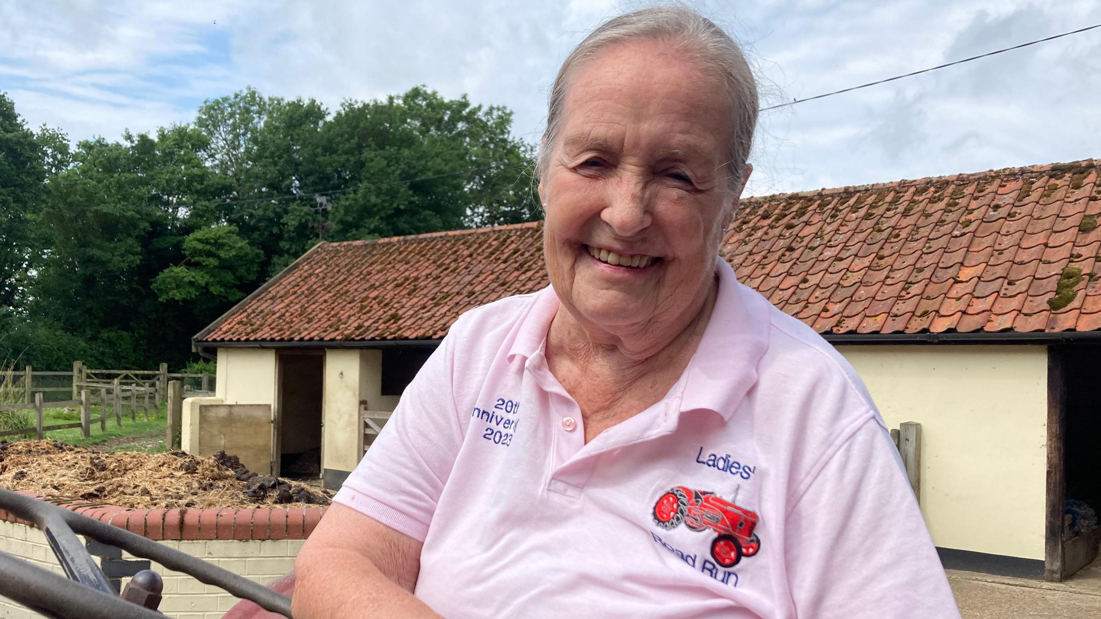 Mrs Chapman is smiling at the camera and wearing her Ladies' Road Run polo shirt. She is sitting at the wheel of a tractor and there are farm buildings and a field behind her.