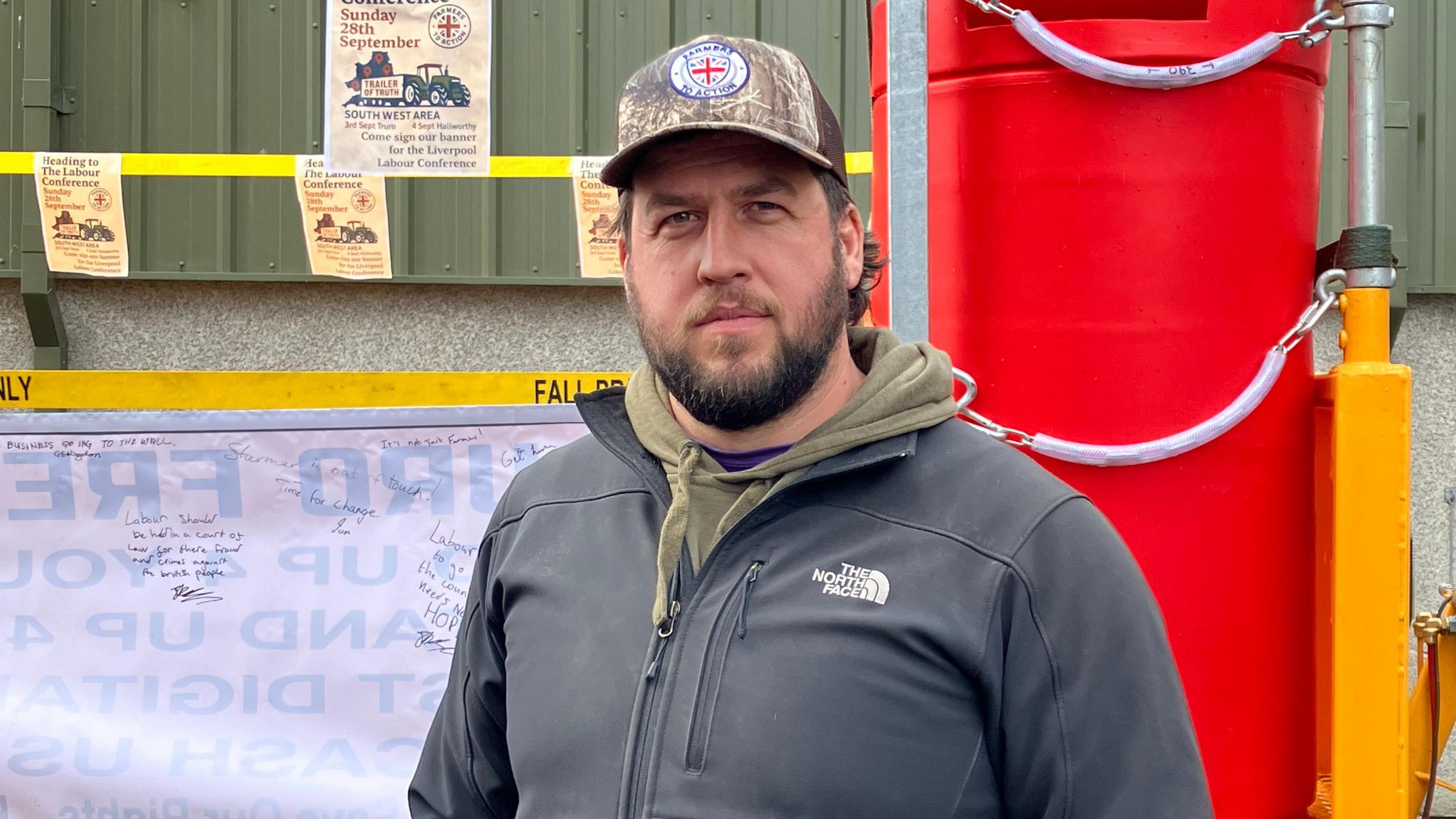 A man wearing a dark grey jacket and a brown hoodie and baseball cap standing in front of a yellow trailer and banner with messages on it.