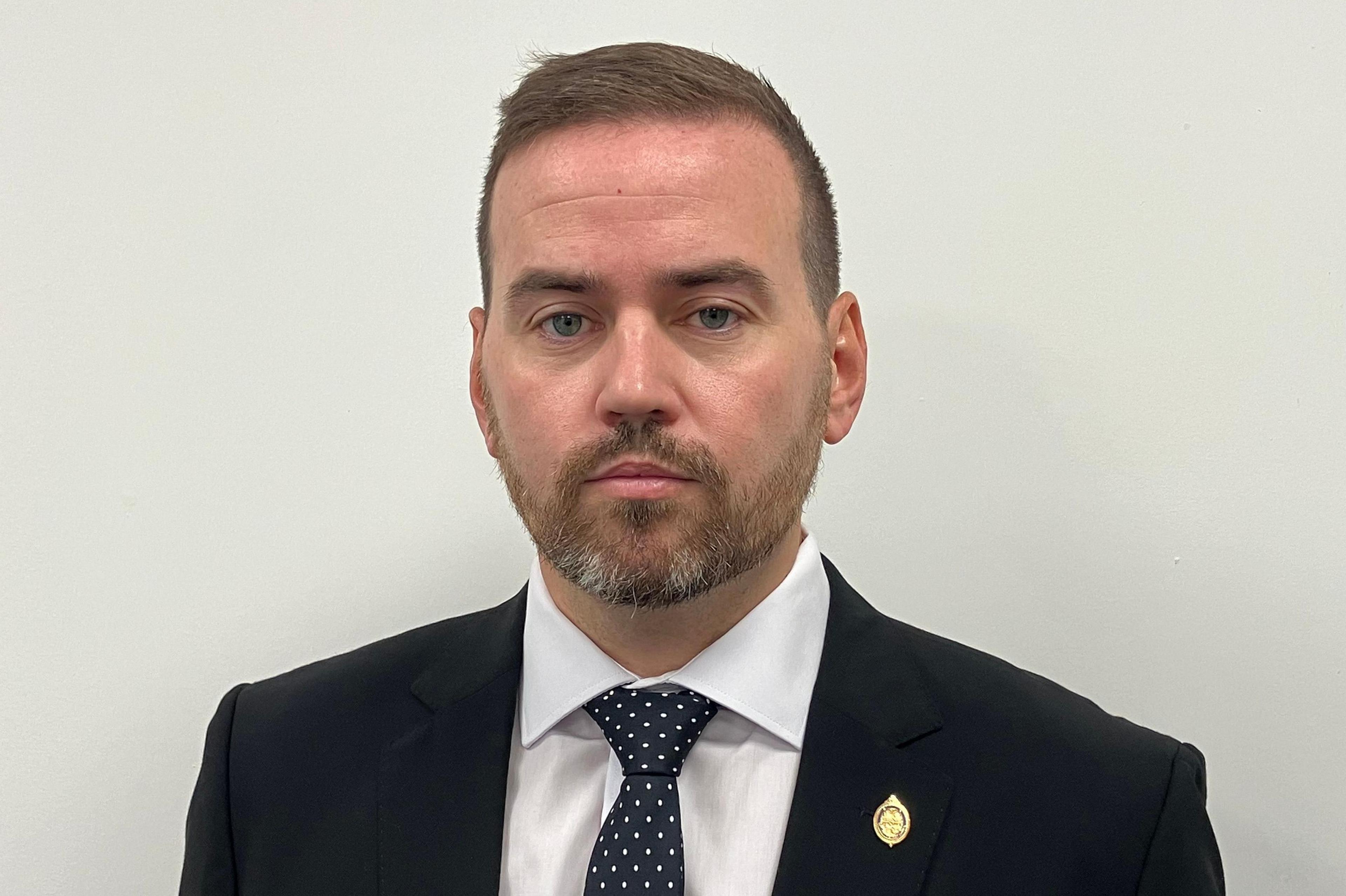 Gavin Callaghan, appearing bearded, with short brown hair, and a black or navy blue suit. He is looking at the camera, not smiling, with an off-white painted wall behind him.