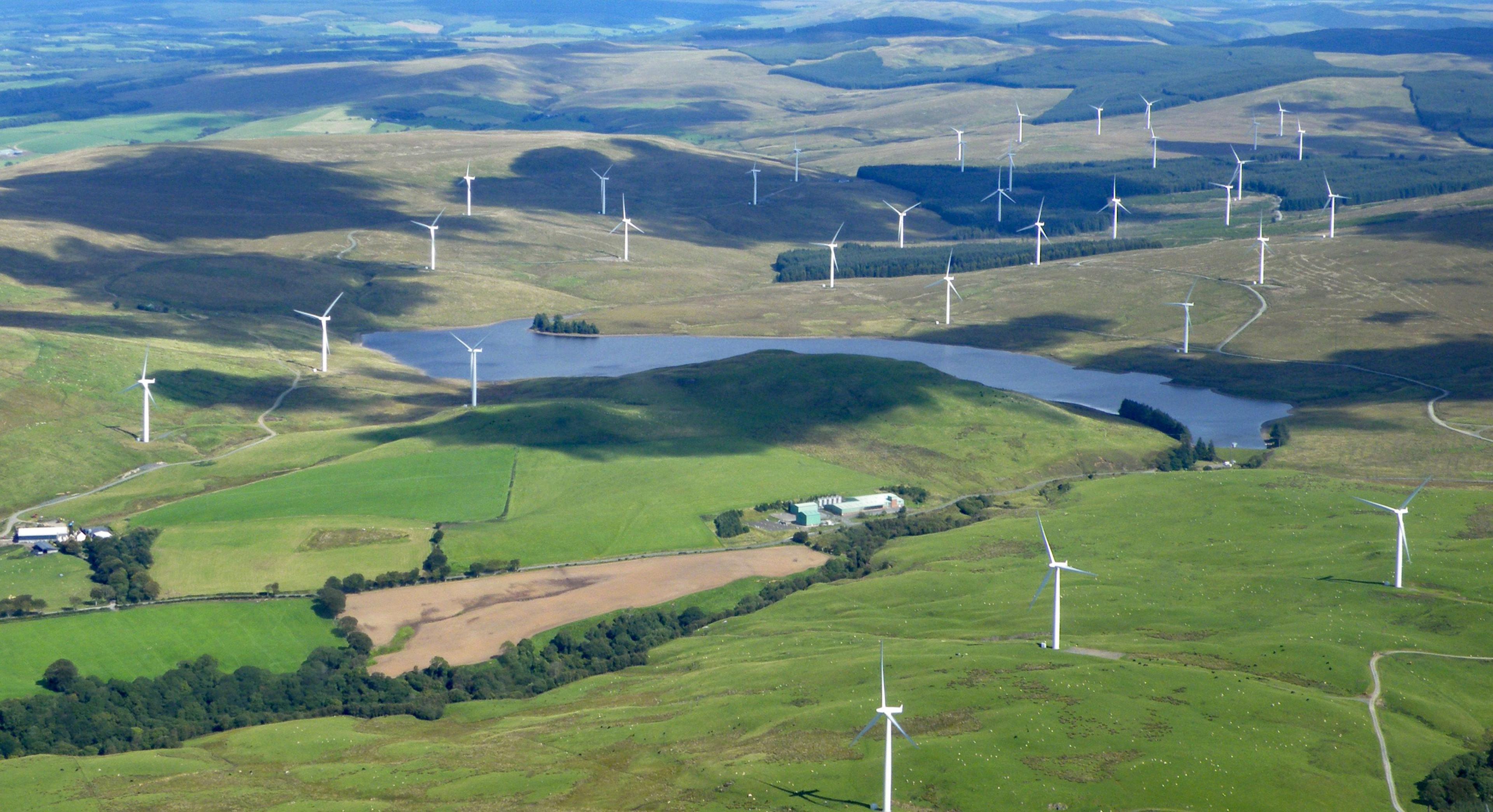 Aerial image of a reservoir surrounded by grass covered hills with wind turbines scattered around the area far into the distance
