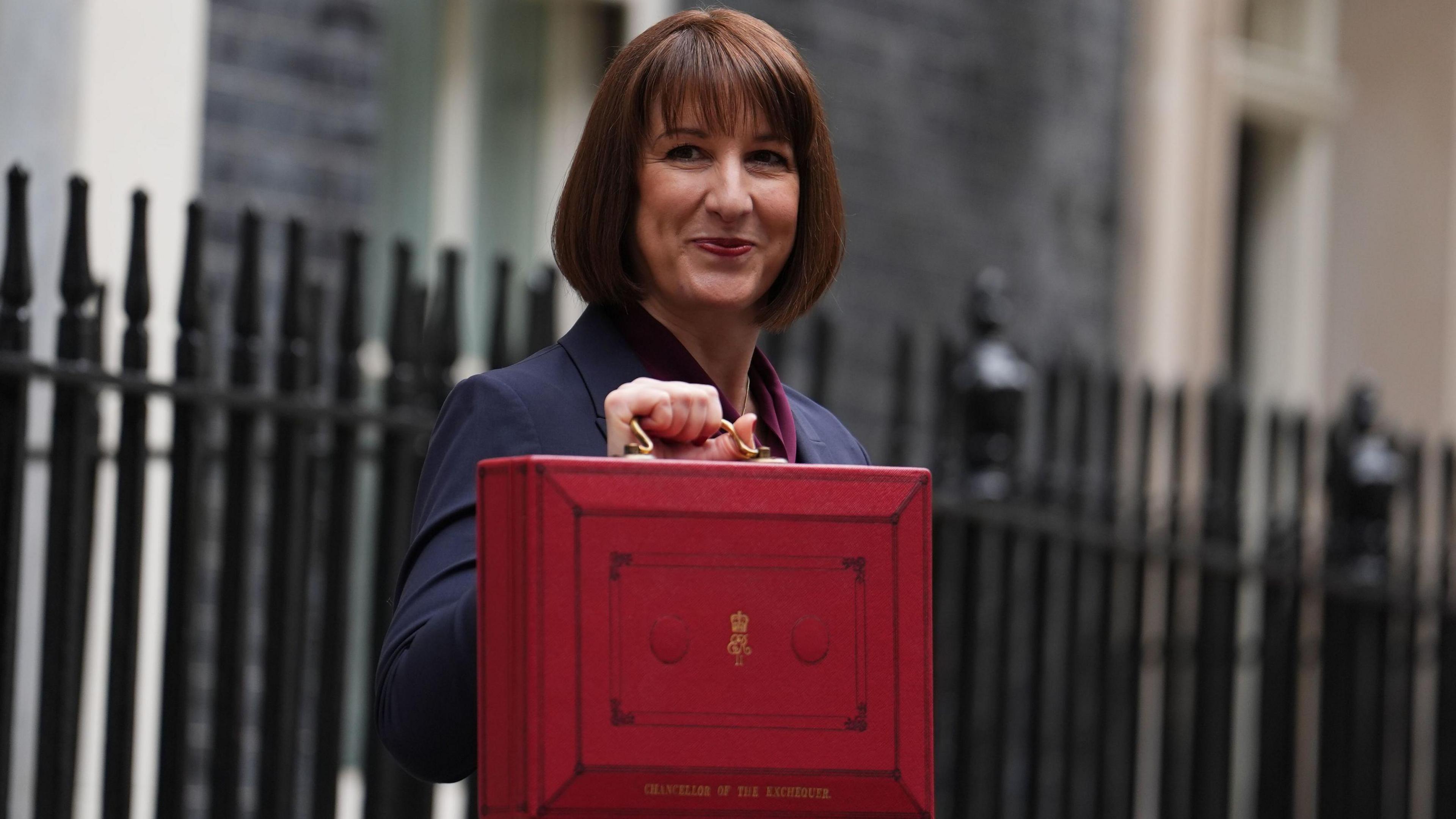 The chancellor, Rachel Reeves, standing in Downing Street holding the Chancellor of the Exchequer red box. She is wearing a dark suit with a red shirt. 