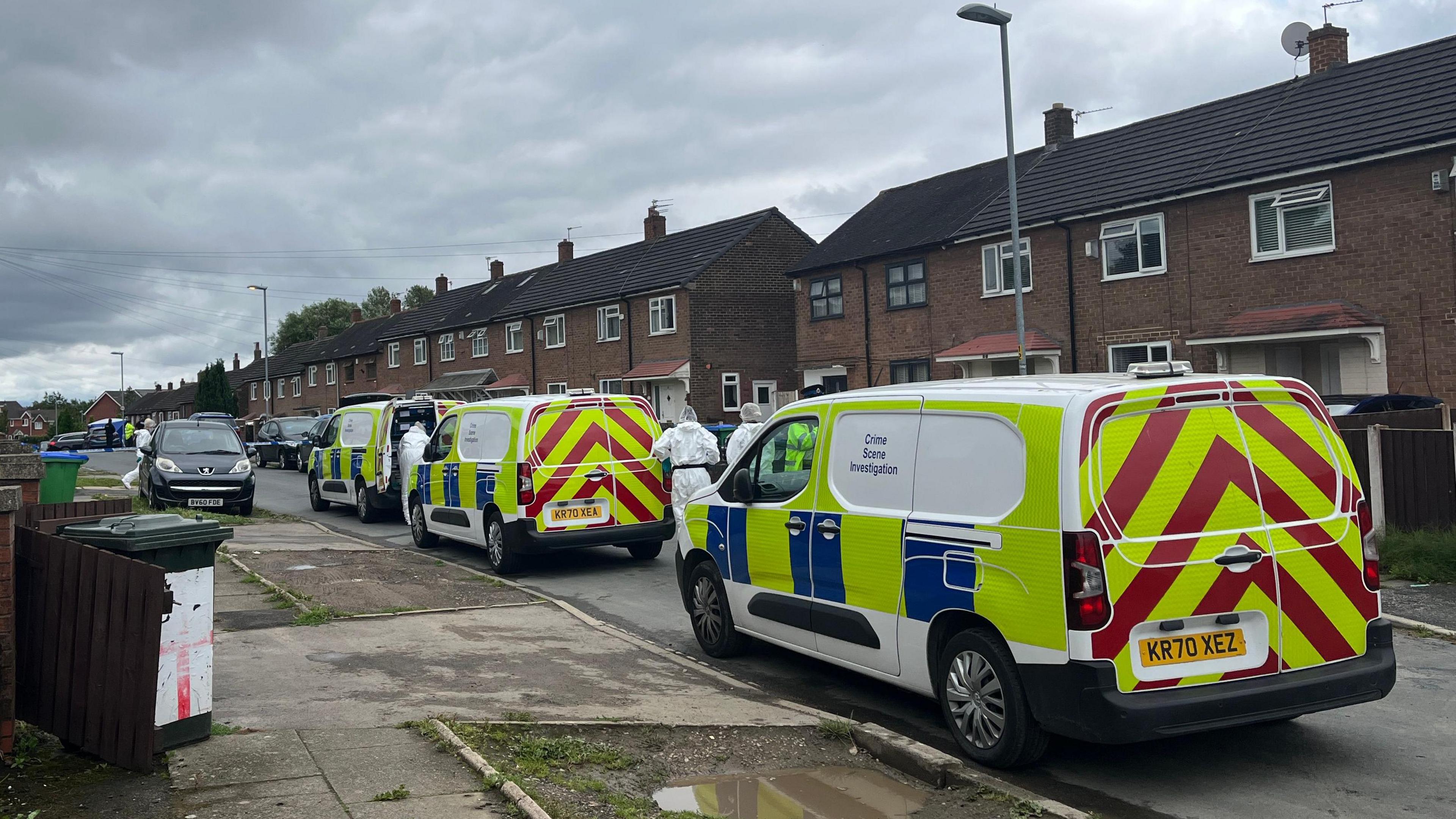 Three police vans are parked on a residential street. Two forensic officers in full white suits and hair nets are walking alongside the vans.