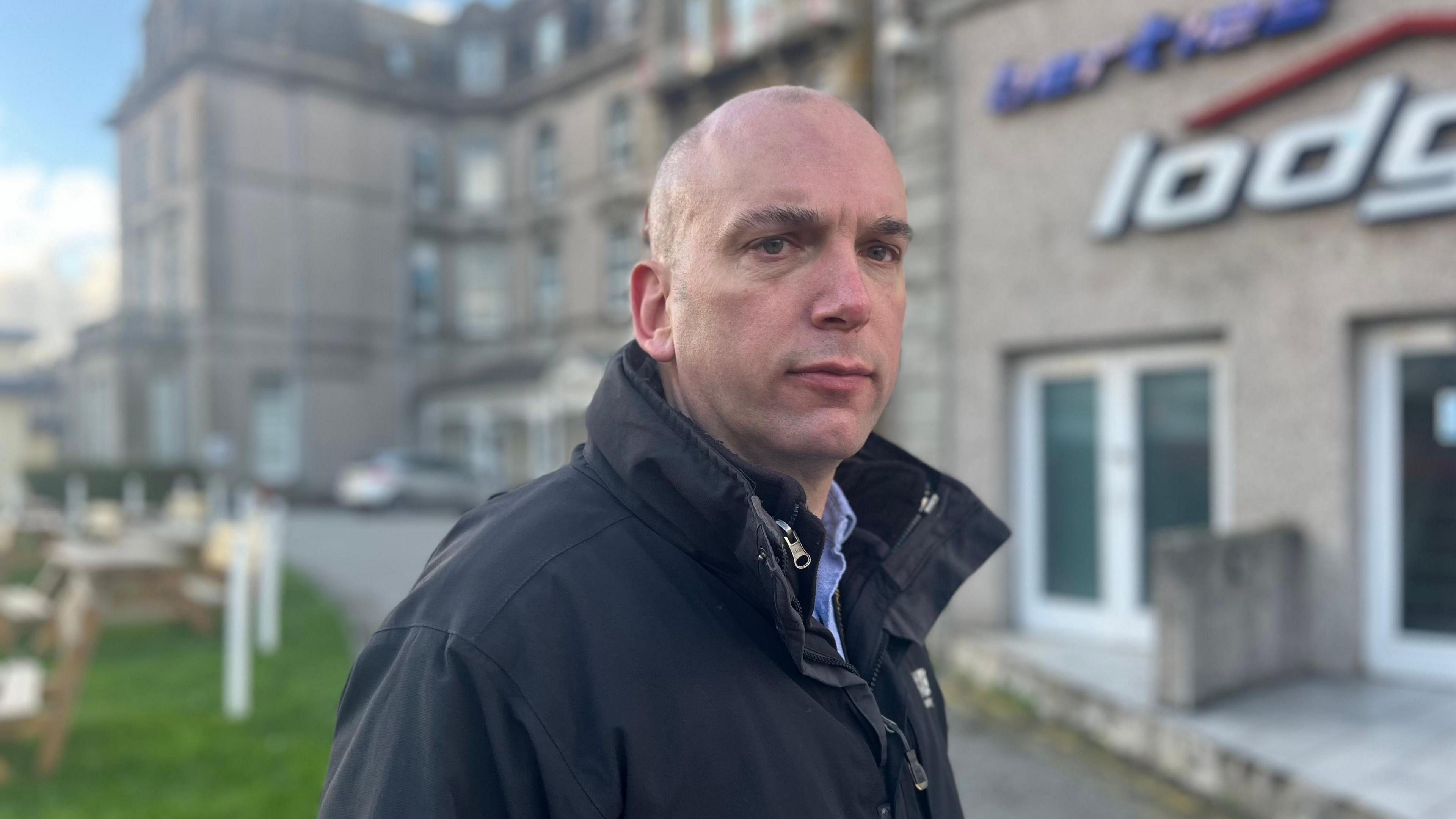 Louis Gardner, Newquay Central member for Cornwall Council, stands looking into the camera, wearing a blue coat and with a serious expression 