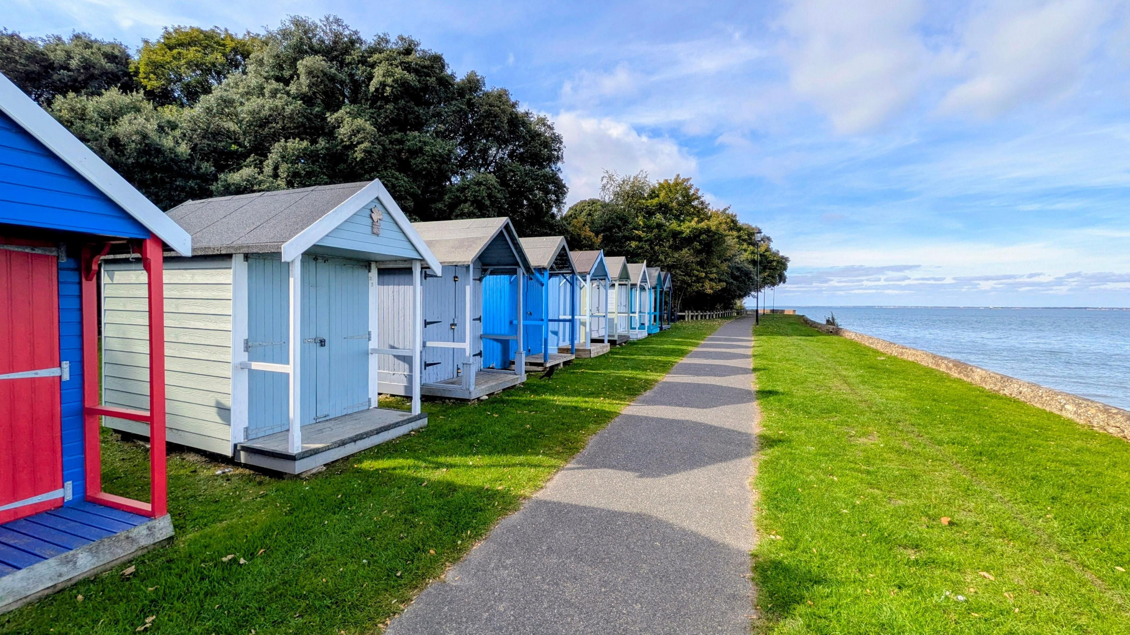 A row of colourful beach huts bask in autumnal sun beside some grass and a tarmac path alongside the sea. the sky is blue with lots of wispy clouds.