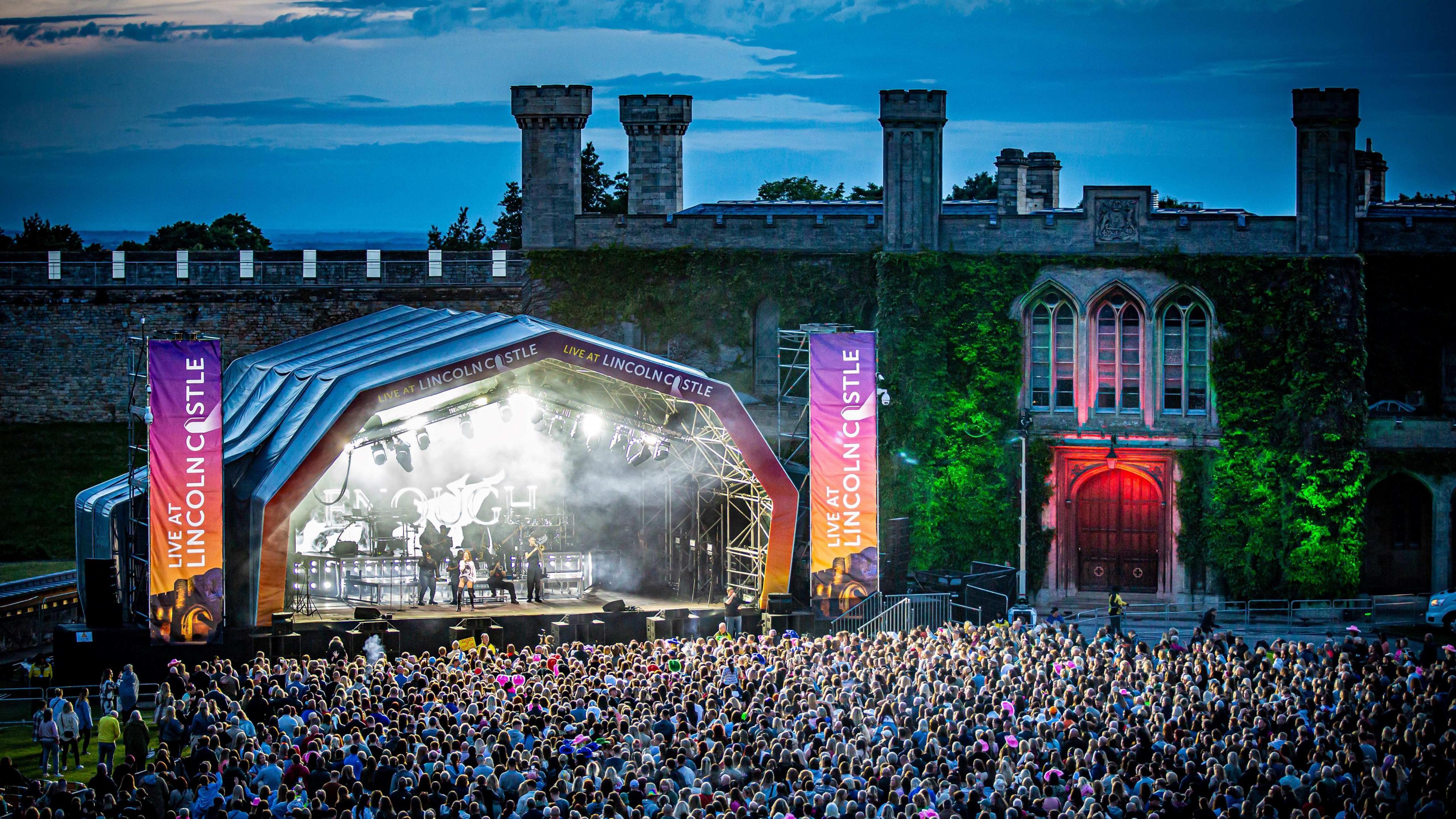 A concert stage with lights and musicians in front of Lincoln Castle with a crowd of people watching on.