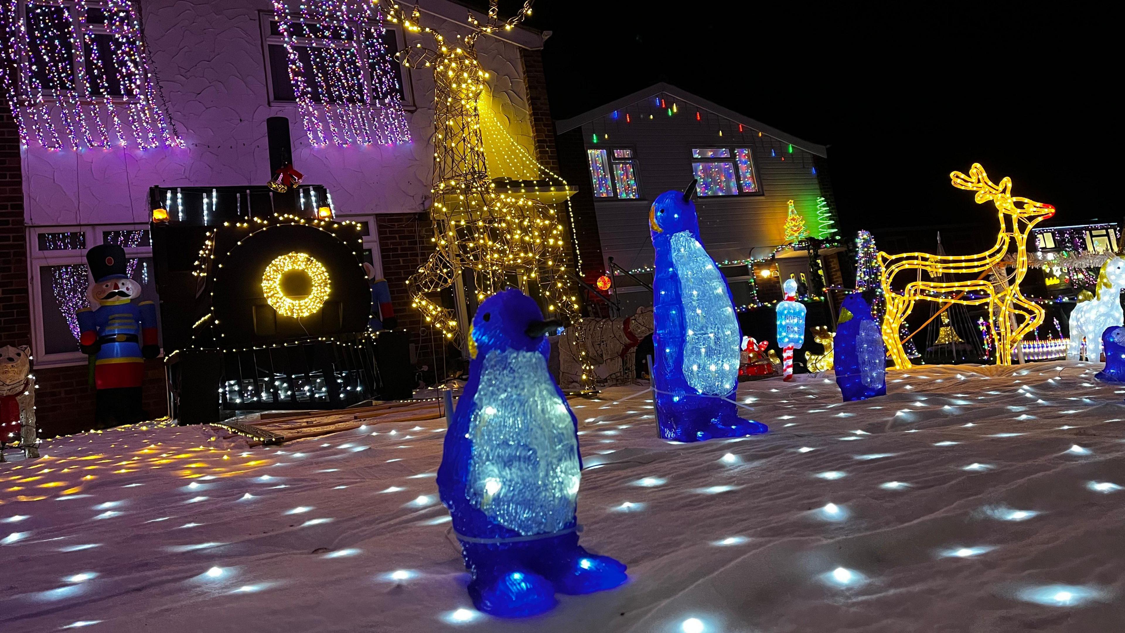 A house's front garden is filled with illuminated penguin figurines, strings of fairy lights, a large decorative train covered in lights and a big light-up reindeer. All of them sit atop a blanket designed to look like snow.