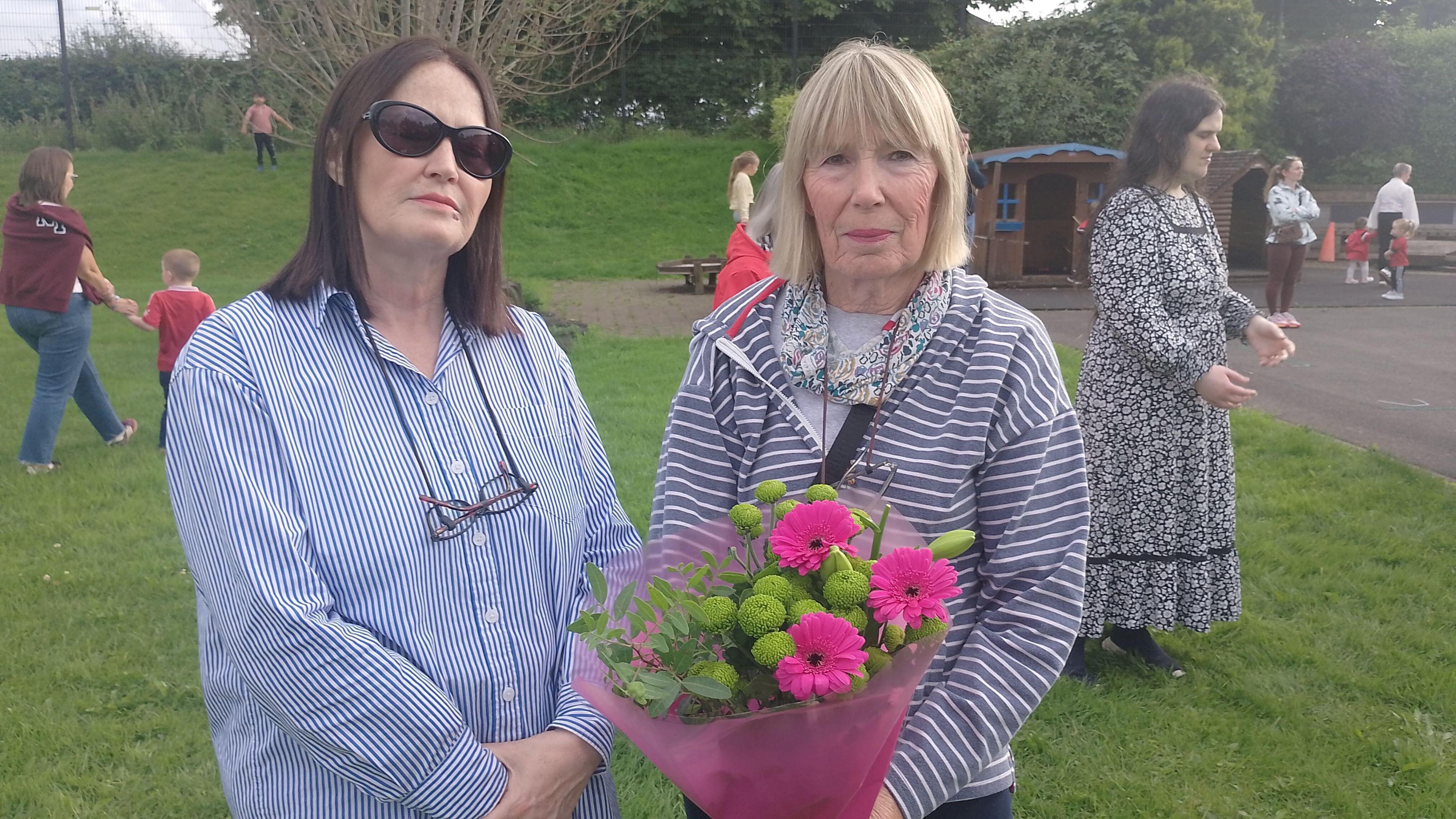 Linda, left, is wearing dark sunglasses. She is wearing a blue and white striped shirt and has shoulder-length brown hair. Jessie, right, is looking at the camera. She has a blonde bob hairstyle and is holding a bunch of pink and green flowers in a pink wrapping. 