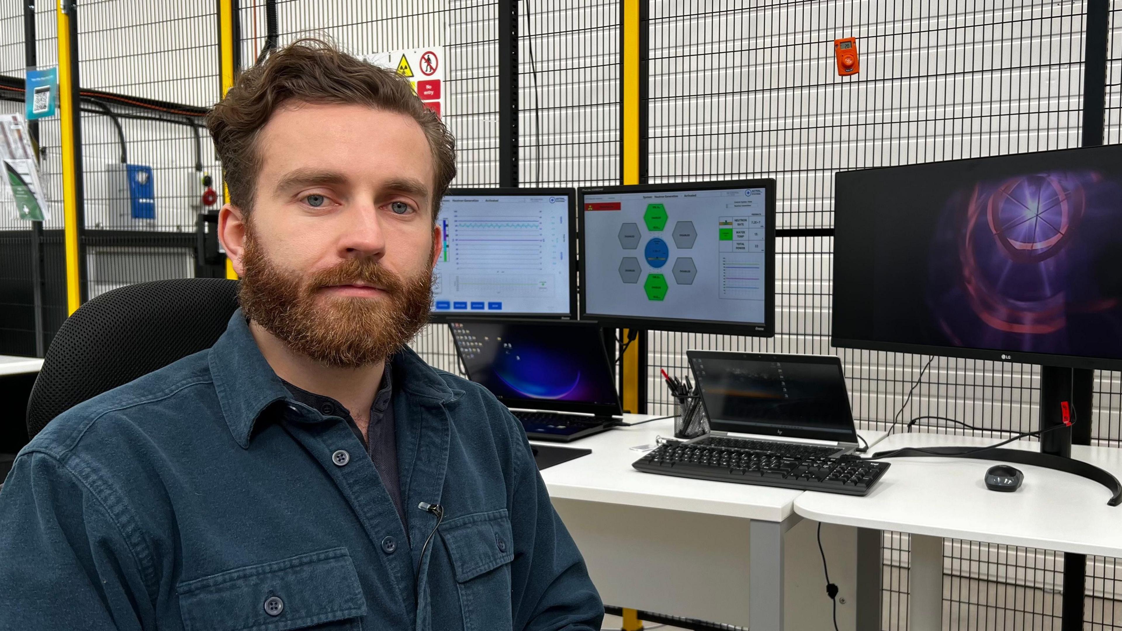 Dr Tom Wallace-Smith sits in his lab, wearing a petrol blue corduroy shirt. He has a brown beard. Behind him, screens show colourful graphics illustrating the technology he has built.