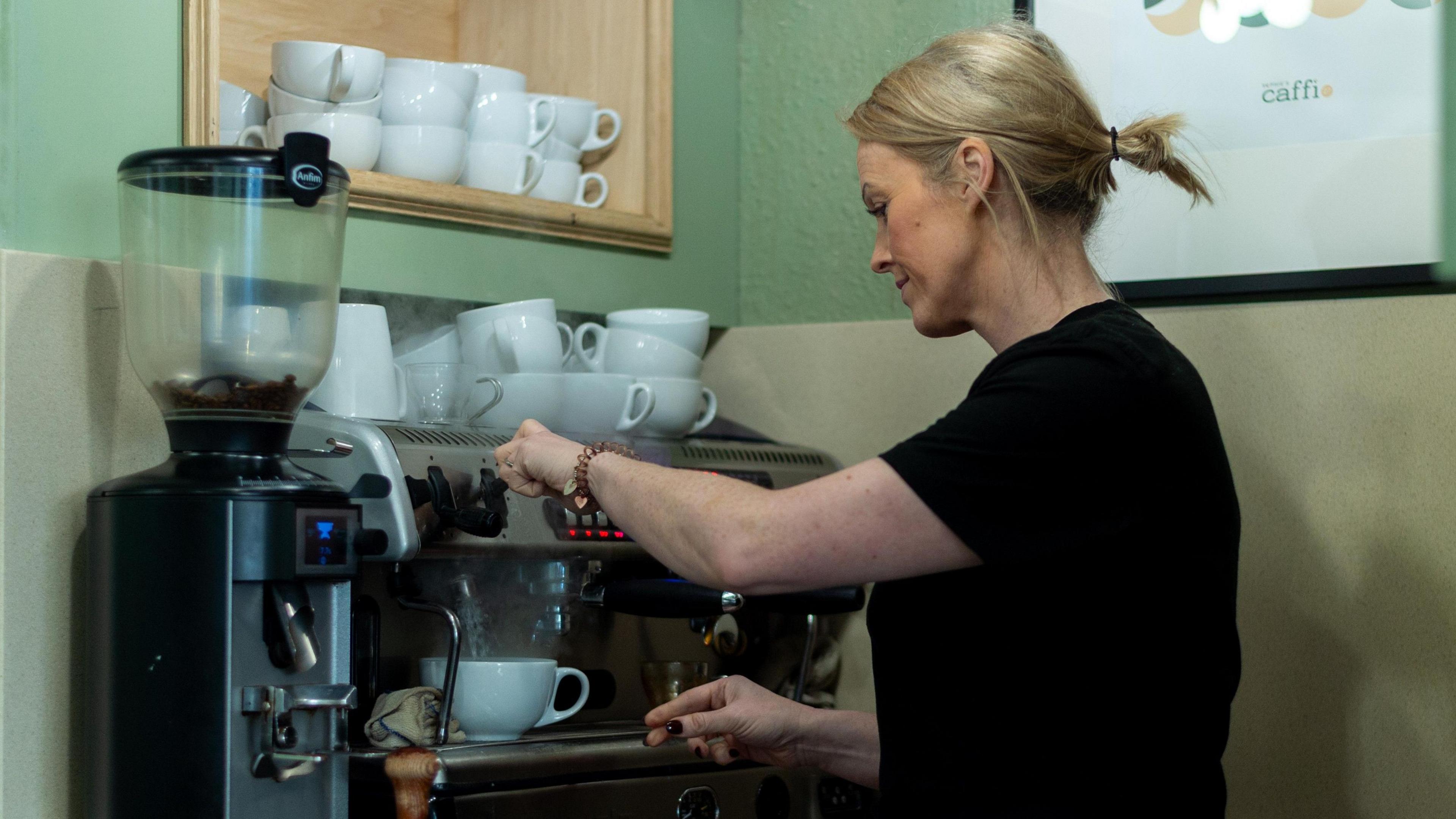 Llun o berchennog caffi Sophie's Caffi 27 yn defnyddio'r peiriant coffi. Mae gan Sara wallt byr melyn ac mae hi'n gwisgo dillad du. Mae hi'n llenwi cwpan gyda dwr poeth. Mae hi'n sefyll o flaen y peiriant, ac mae'r wal y tu ol iddi yn wyrdd.