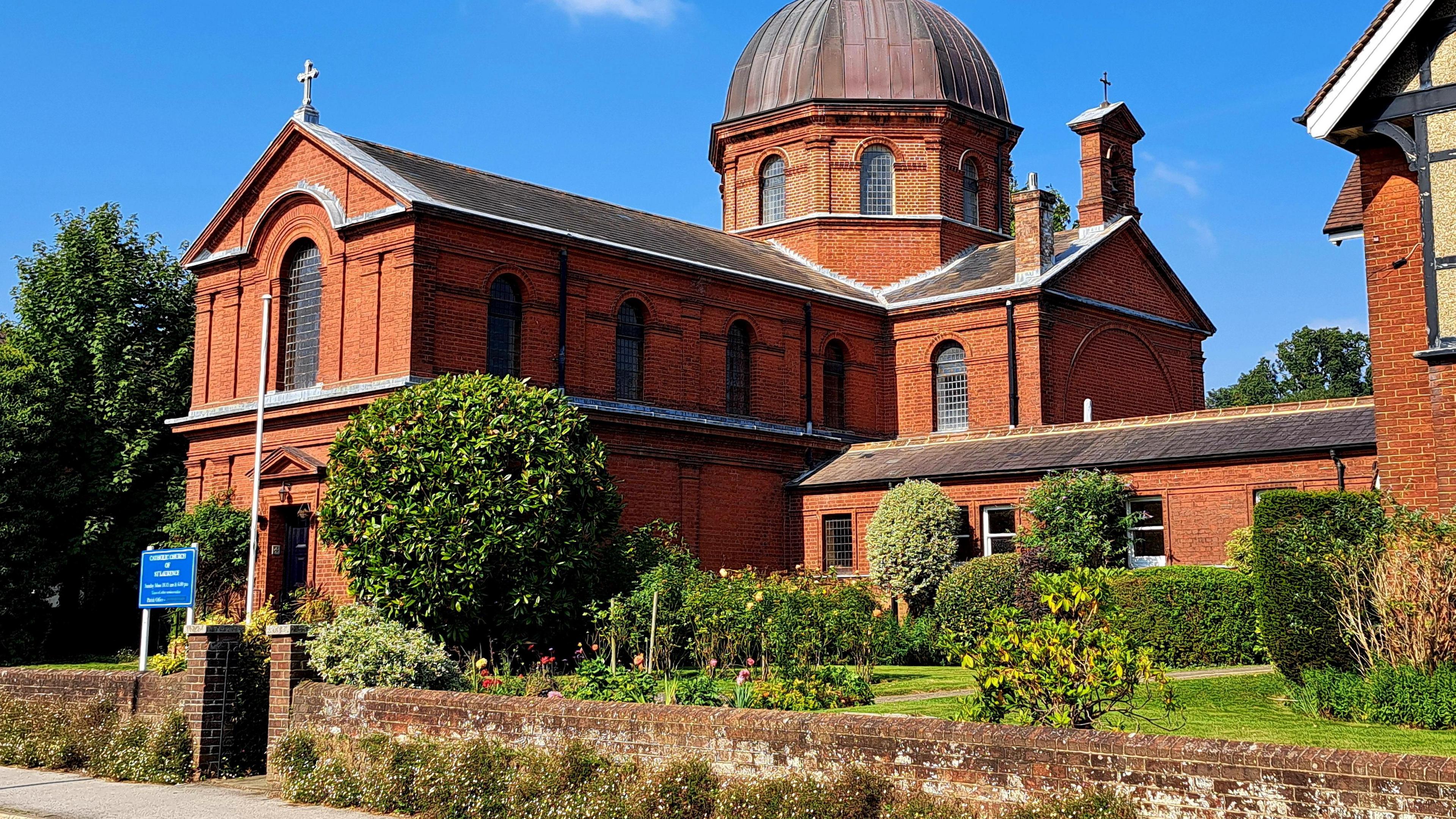 A sunny scene is dominated by a red brick church-type building. It has a dome at the rear and there is a cross at the top of the roof at the front. A garden runs along the building at the front.