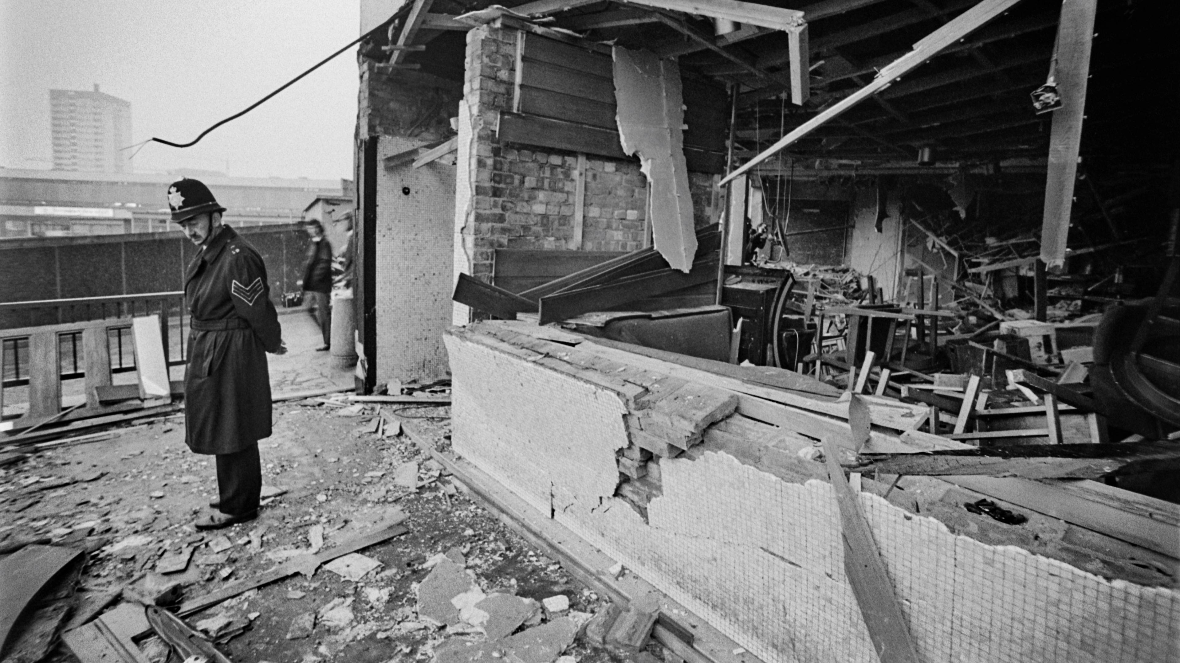 A black and white image showing a policeman standing outside a a building that has been destroyed. The wall is missing and there is rubble and furniture spread around, and bits of the ceiling are dangling down.