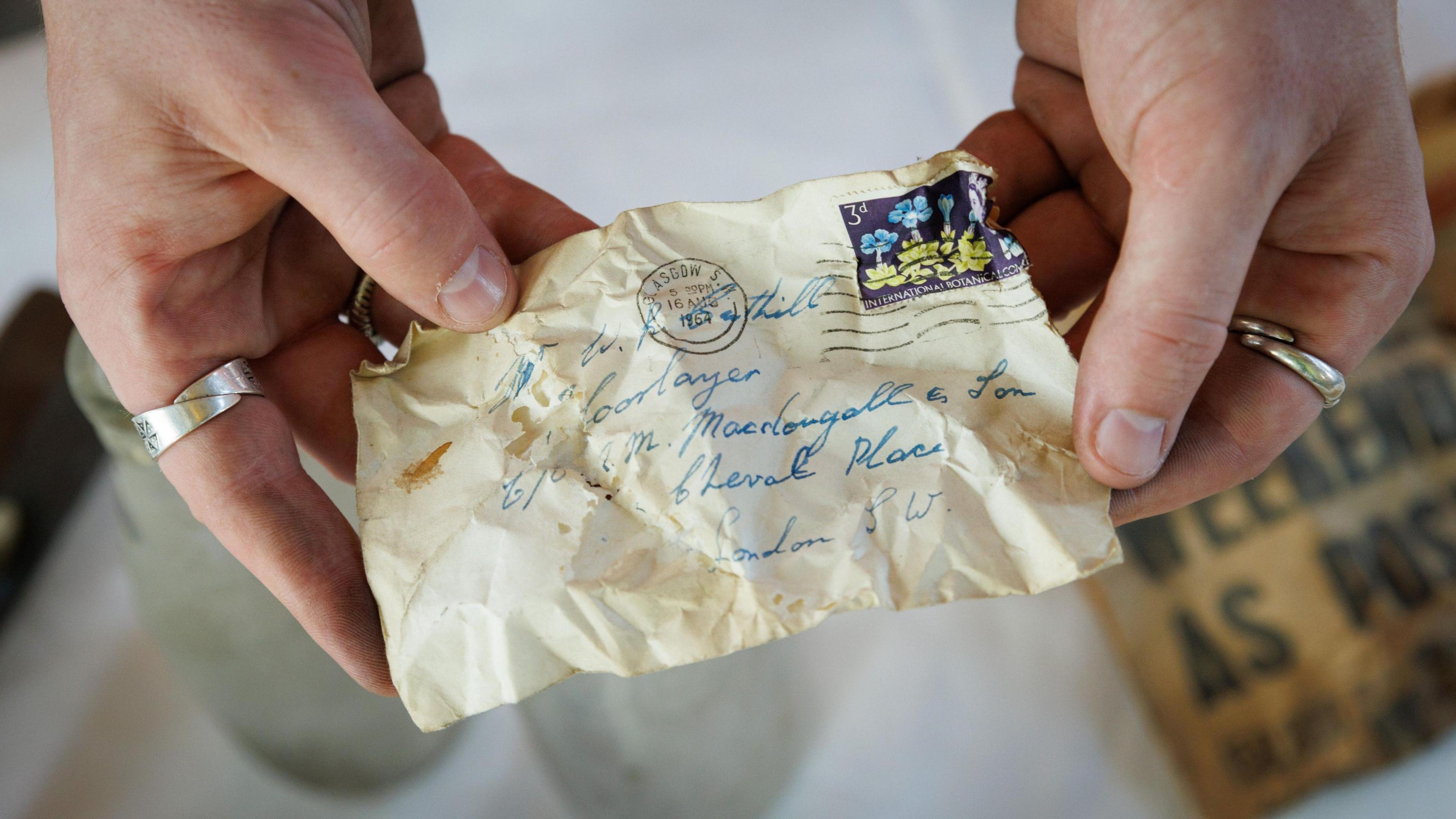 A close-up of a crinkled envlope with a stamp on it addressed to a Mr Cuthill, London SW. There is also a circular stamp with Glasgow 16 August 1964 printed on it. 