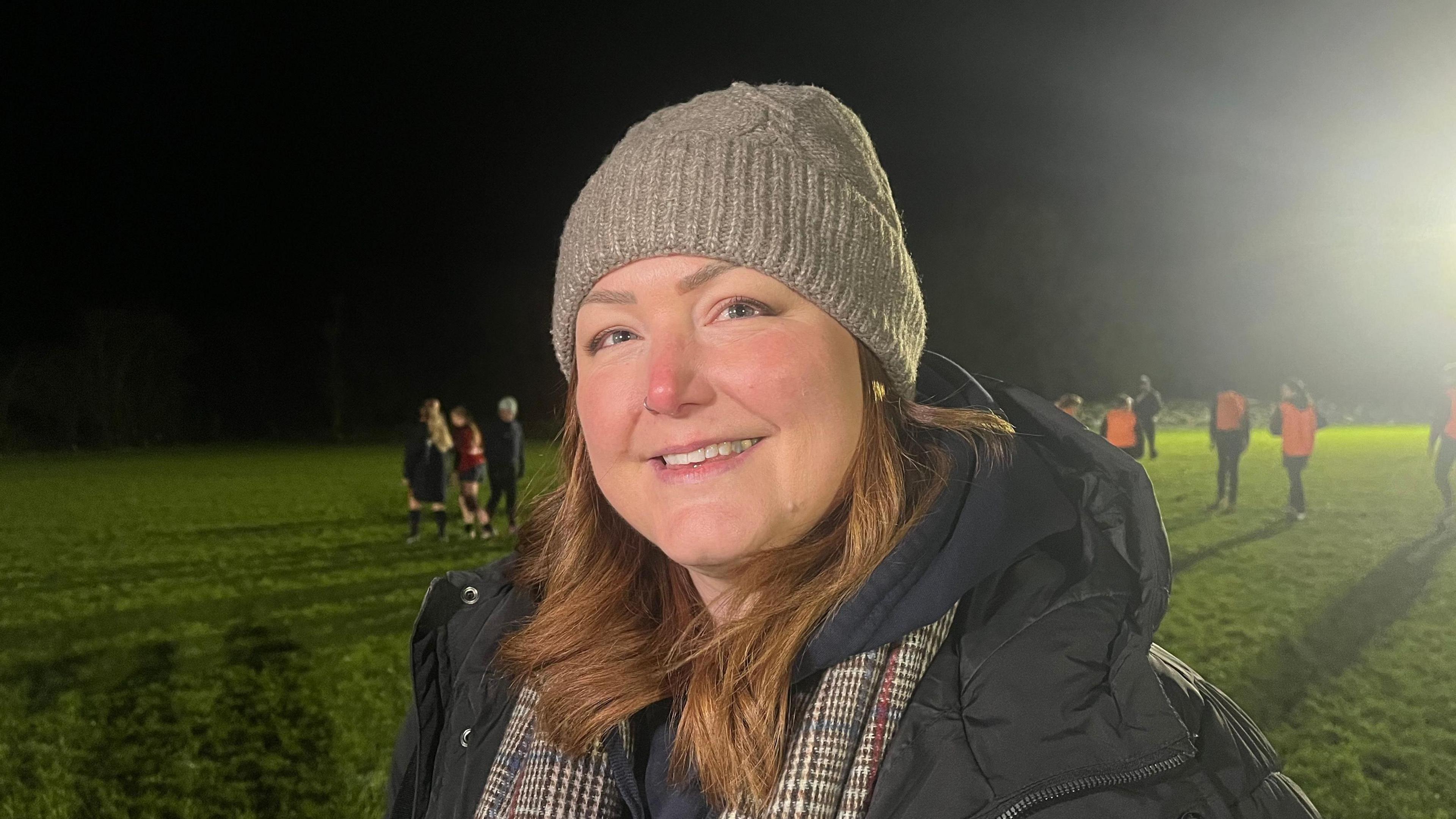 Jessica photographed looking into the camera and smiling. She's wearing a black winter coat, a black hoodie, a brown checked scarf, and a light brown woollen hat. She's stood on a flood-lit rugby pitch, which has green grass, a dark night sky, and is bordered by trees.