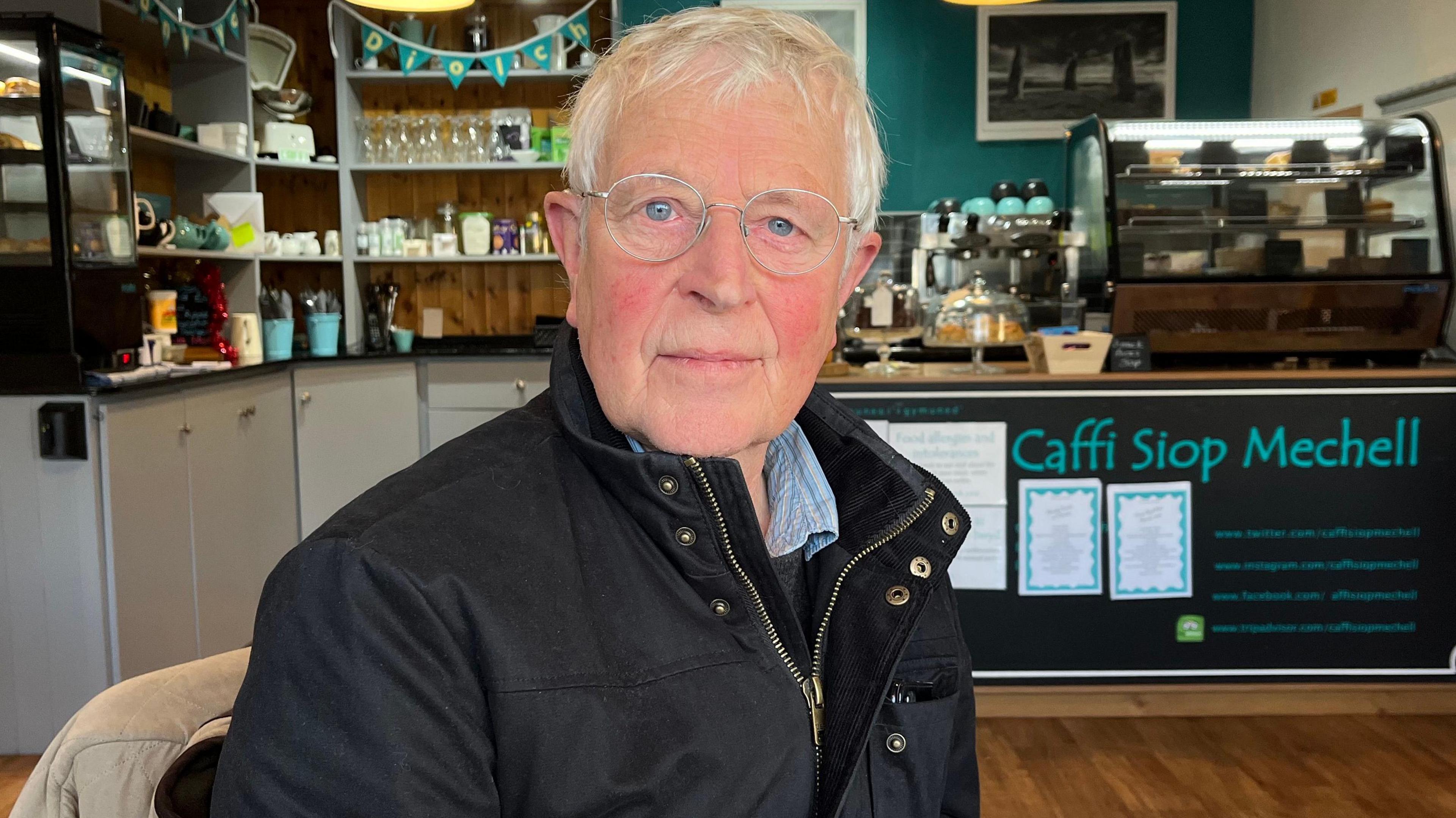 Llun o Jac yn eistedd mewn caffi ac yn edrych ar y camera. Mae ganddo wallt llwyd, llygaid glas a sbectol crwn. Mae'n gwisgo cot du. Tu ôl iddo mae prif gownter y caffi.